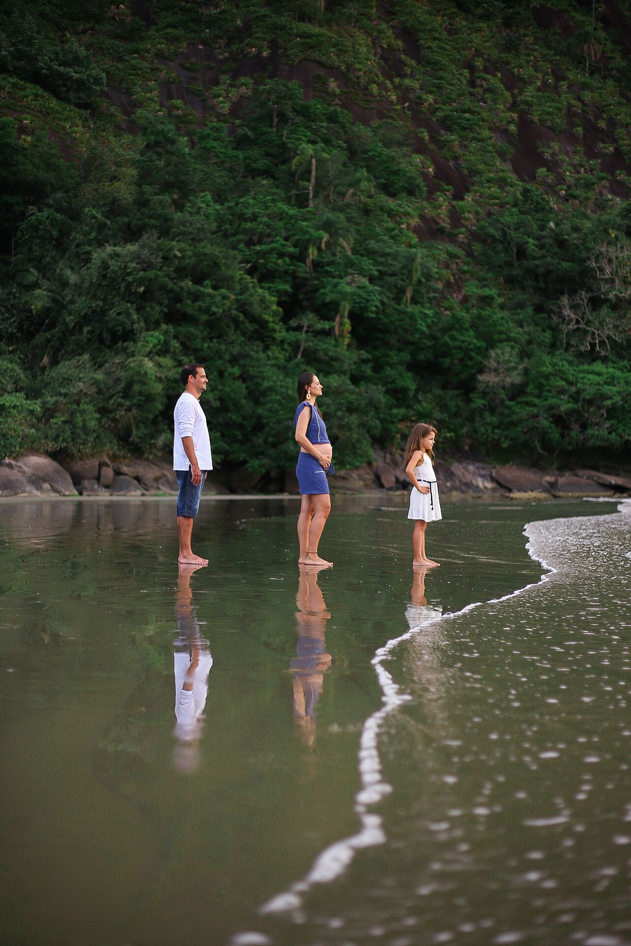Three people standing in shallow waters at beach edge with lush green forest backdrop and gentle waves reflecting their silhouettes.
