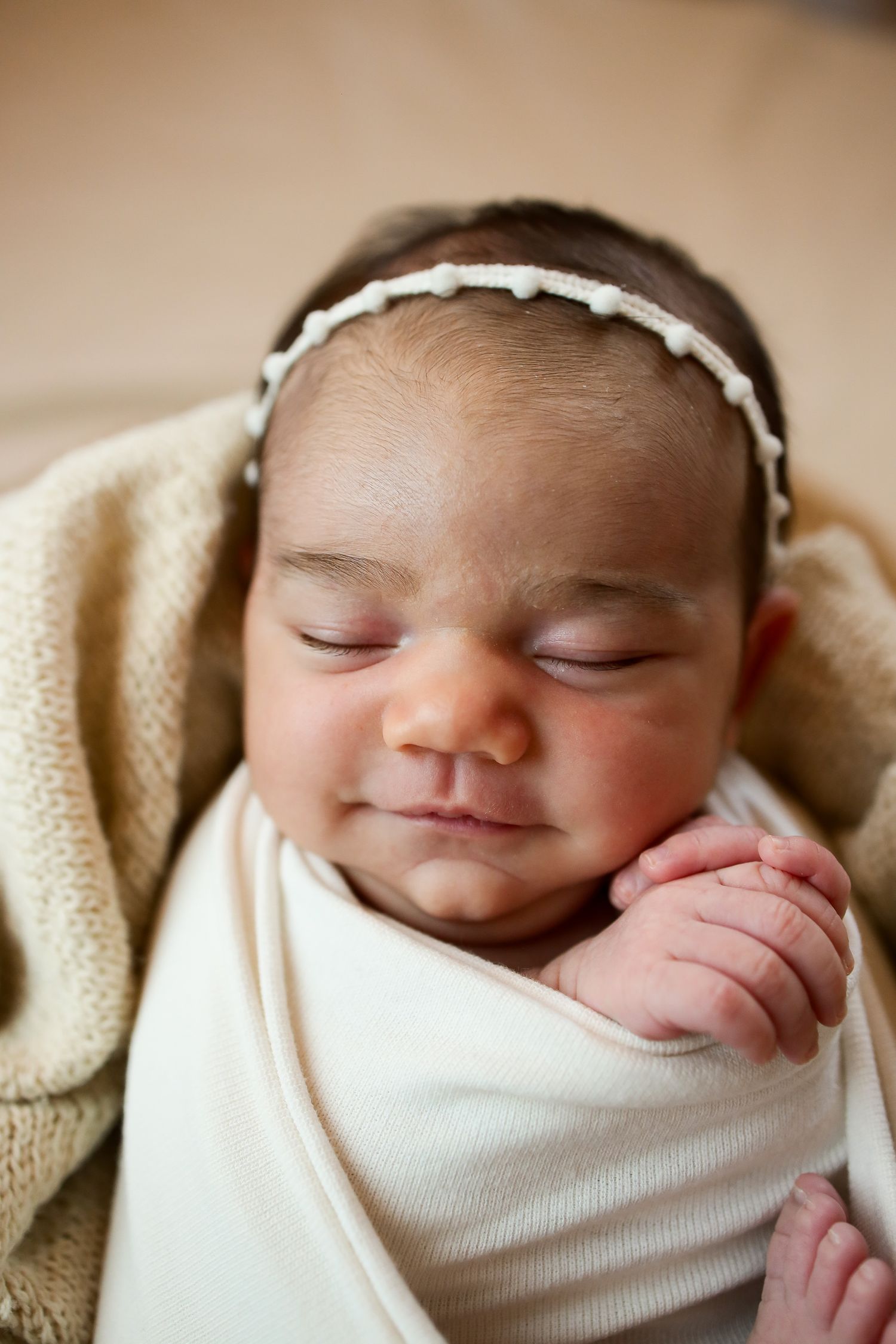 A sleeping newborn wrapped in a white swaddle and wearing a delicate white headband rests peacefully on a beige blanket.