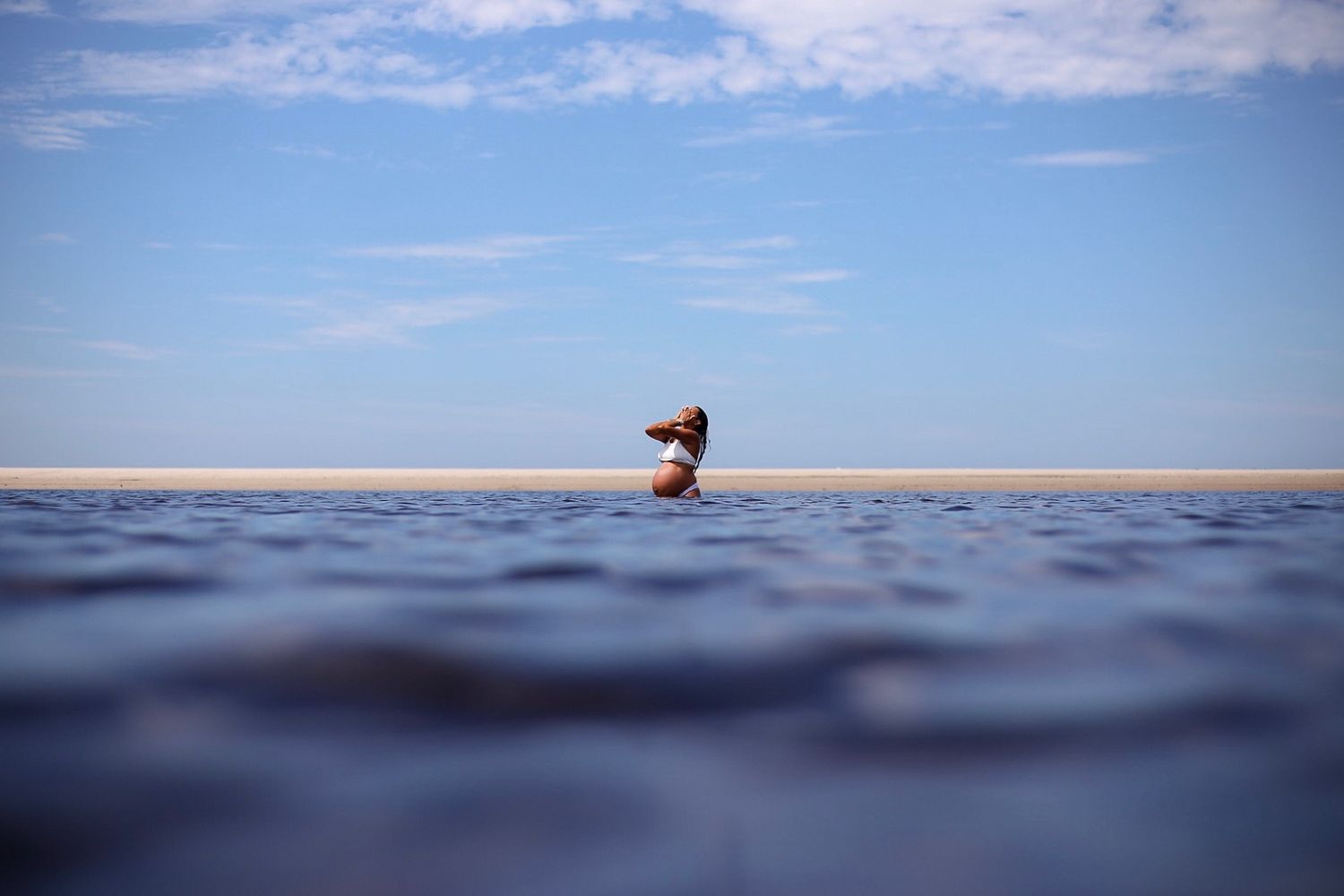 A lone surfer sits on their surfboard in calm ocean waters under a blue sky with wispy clouds.