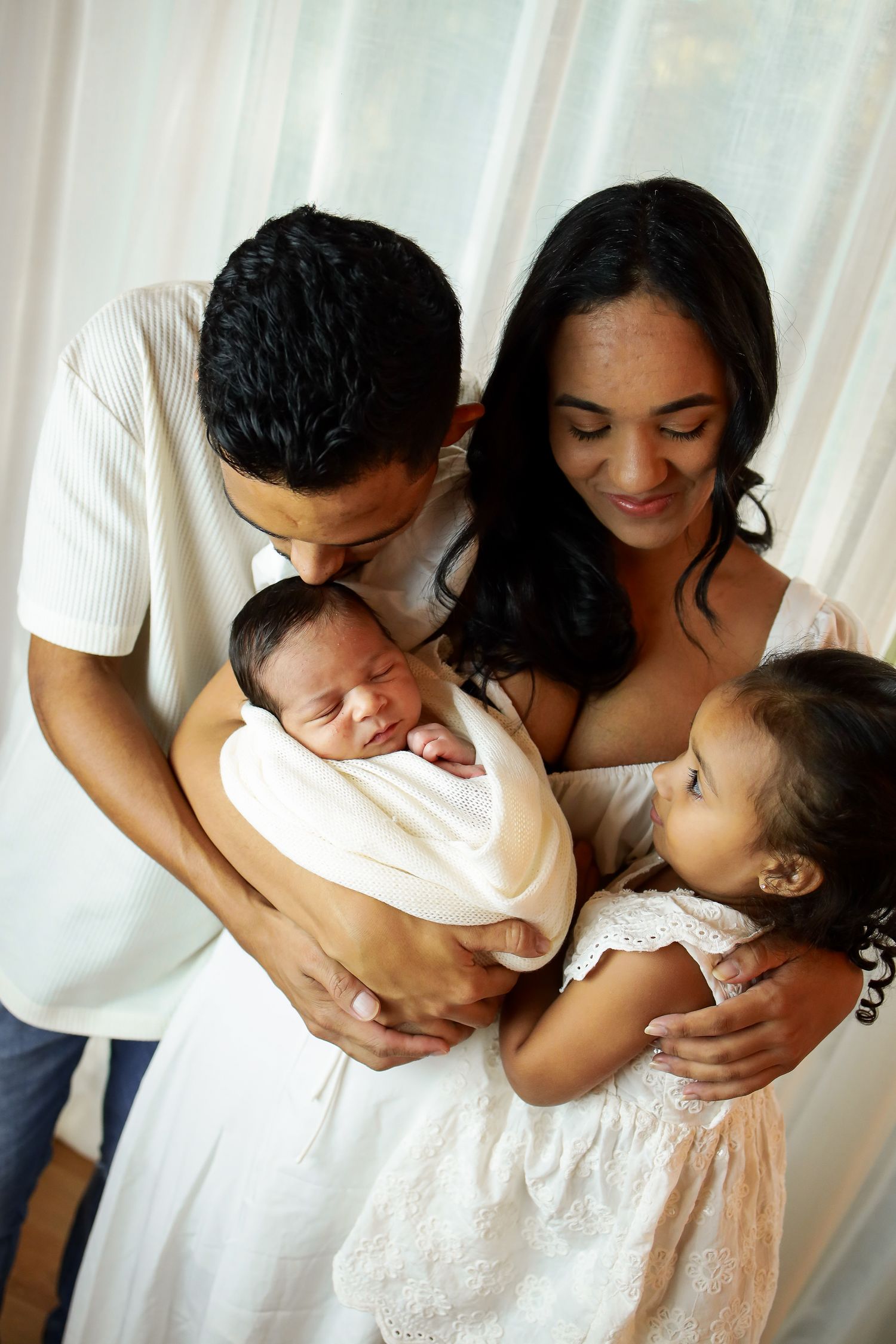 A loving family shares a tender moment together with their newborn baby and toddler daughter in white attire.