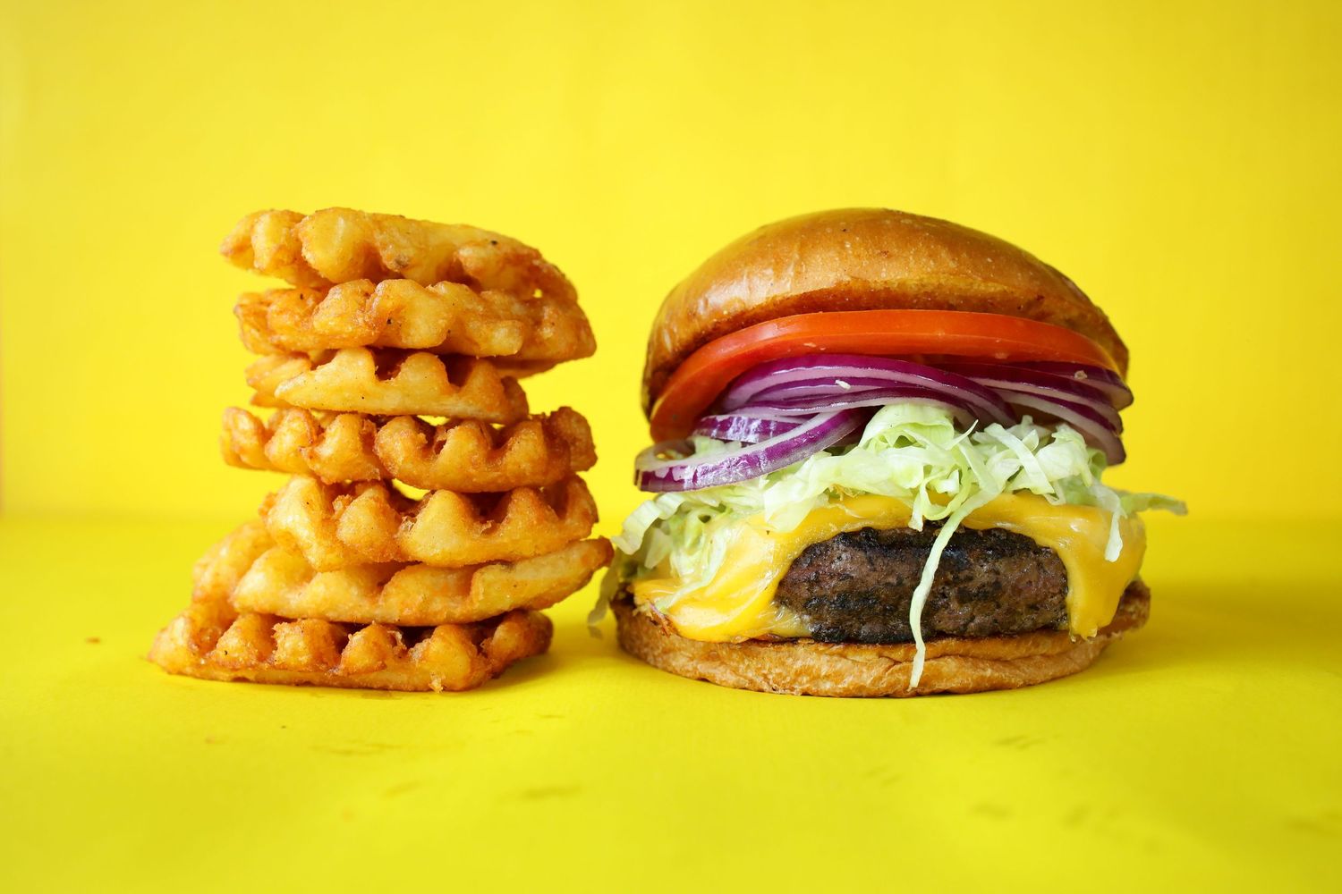 Juicy cheeseburger with lettuce and onions served alongside a stack of crispy waffle fries on yellow background.
