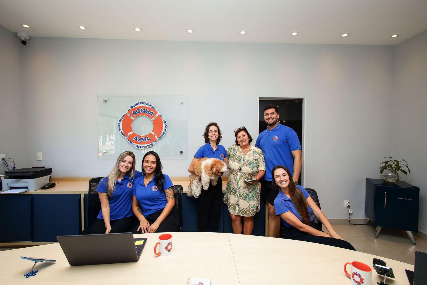 A group of staff members wearing blue shirts gather around a desk in a bright office with a life preserver logo on the wall.