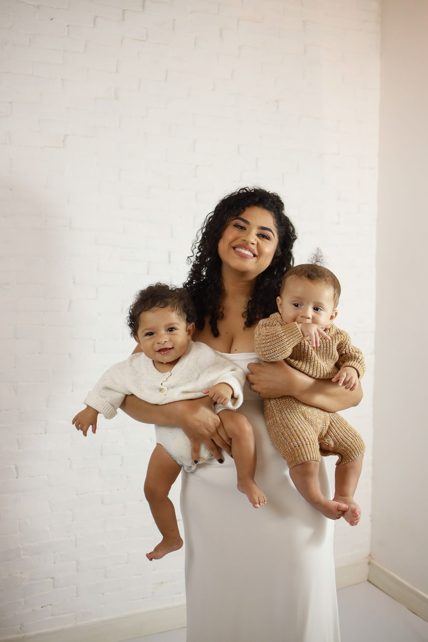 Parent in white dress holding two playful young children against a light-colored wall.