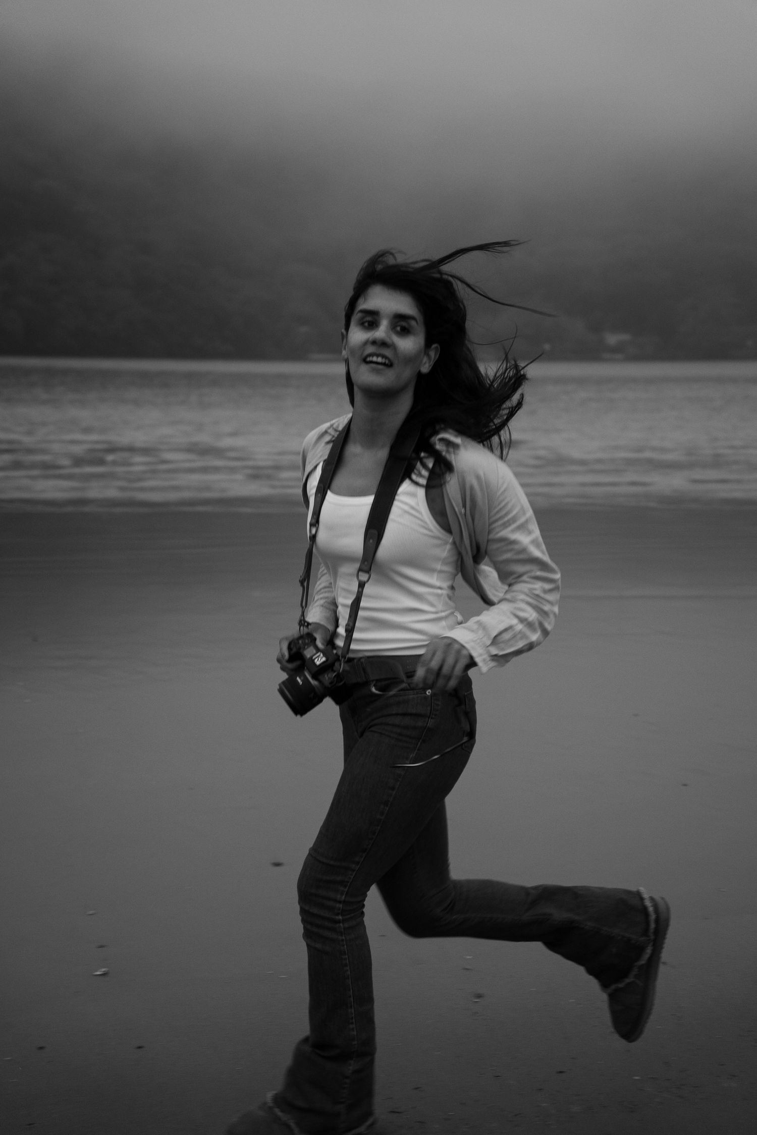 A photographer runs playfully on a foggy beach with a camera around their neck.