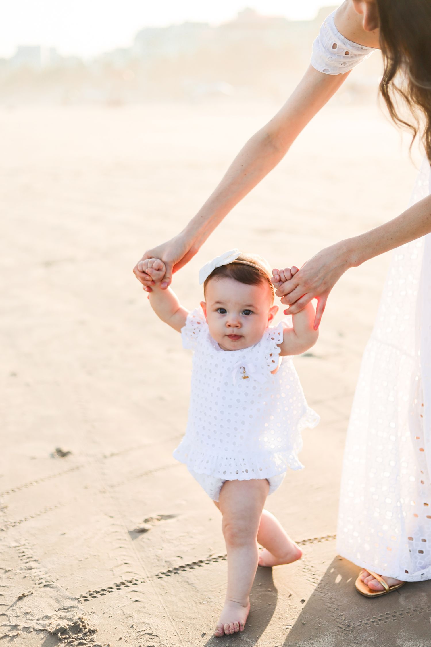 Baby in white eyelet dress takes first steps on beach while being supported by parent's hands during sunset.