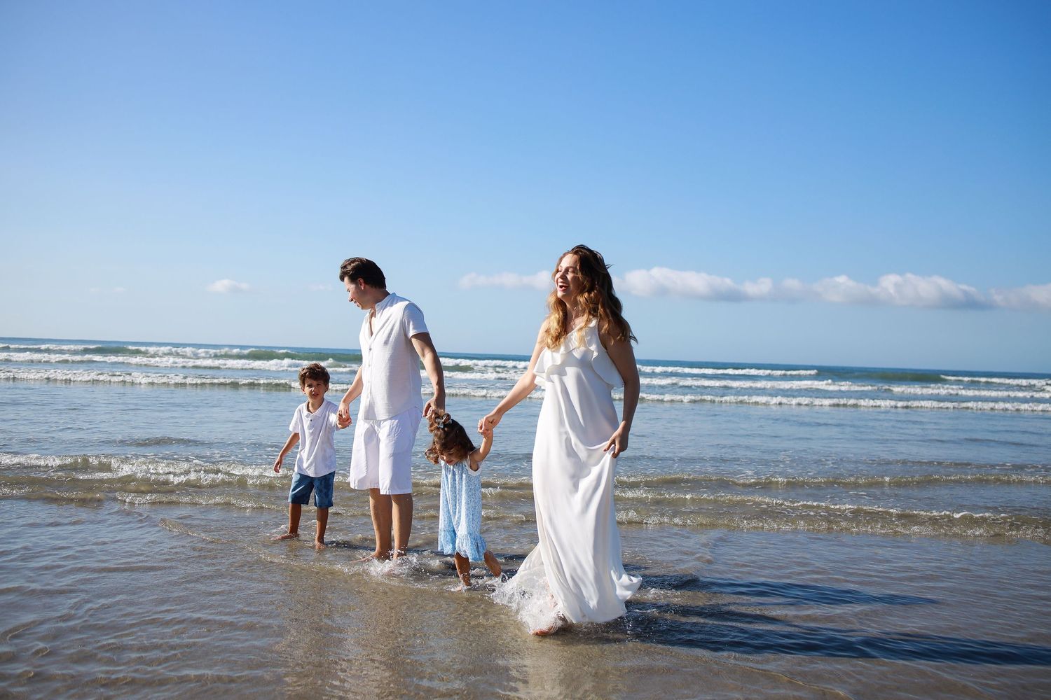 Group in white clothing walking along scenic beach shore with gentle waves and blue sky in background.