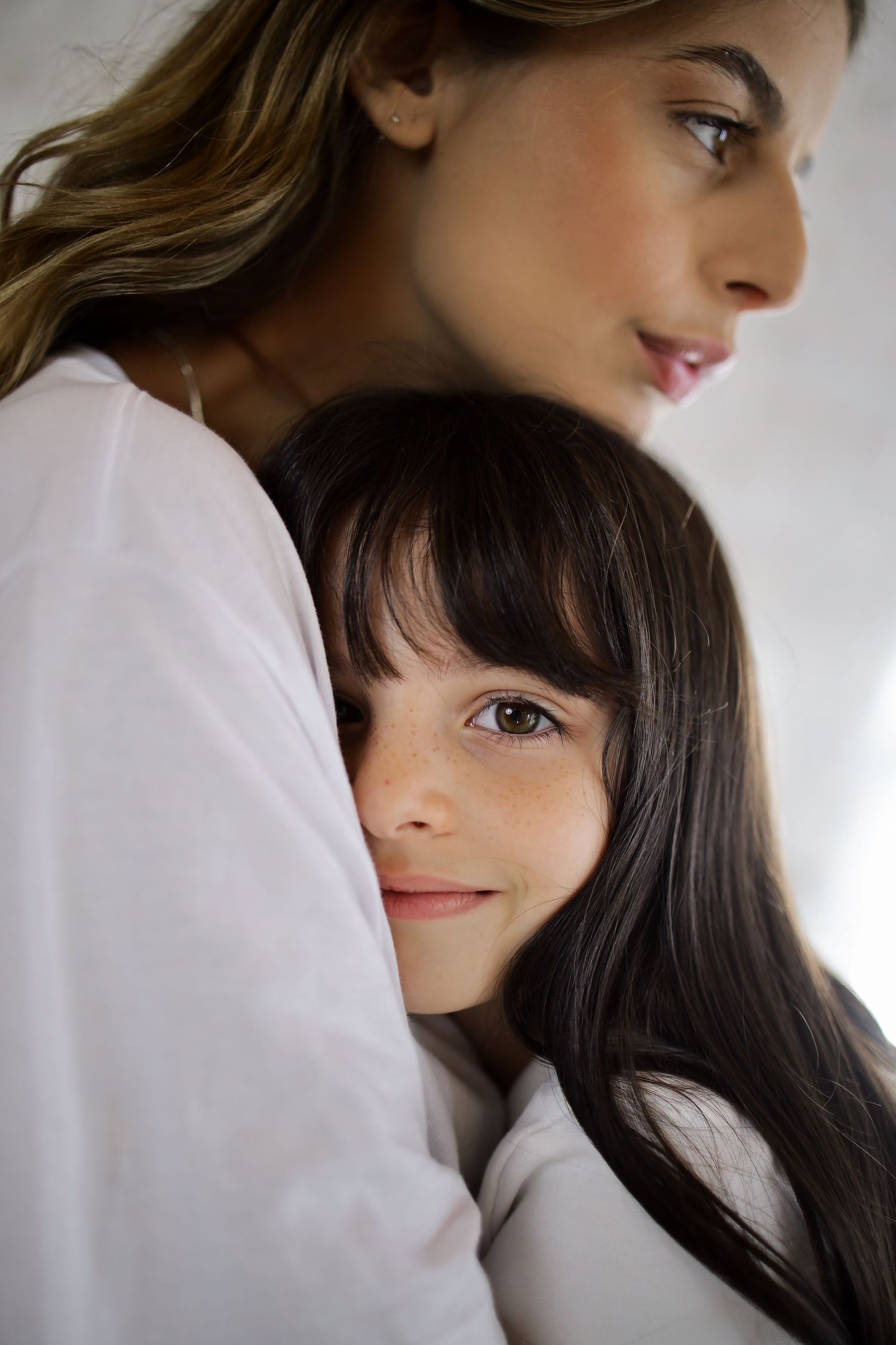 A young girl with dark hair hugs a smiling woman in white, both sharing a warm, tender moment together.