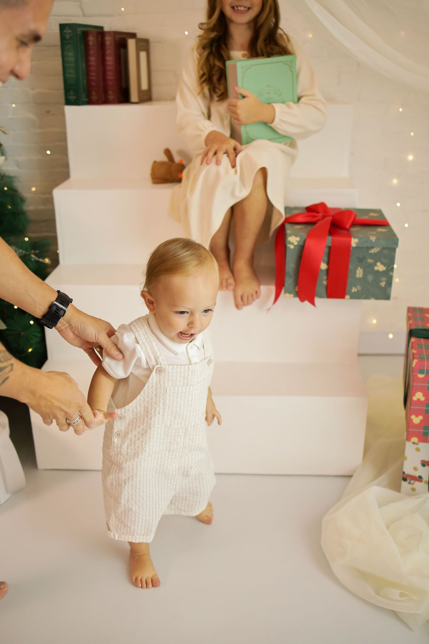 Baby in white romper takes first steps on white stairs during Christmas with wrapped presents in background.