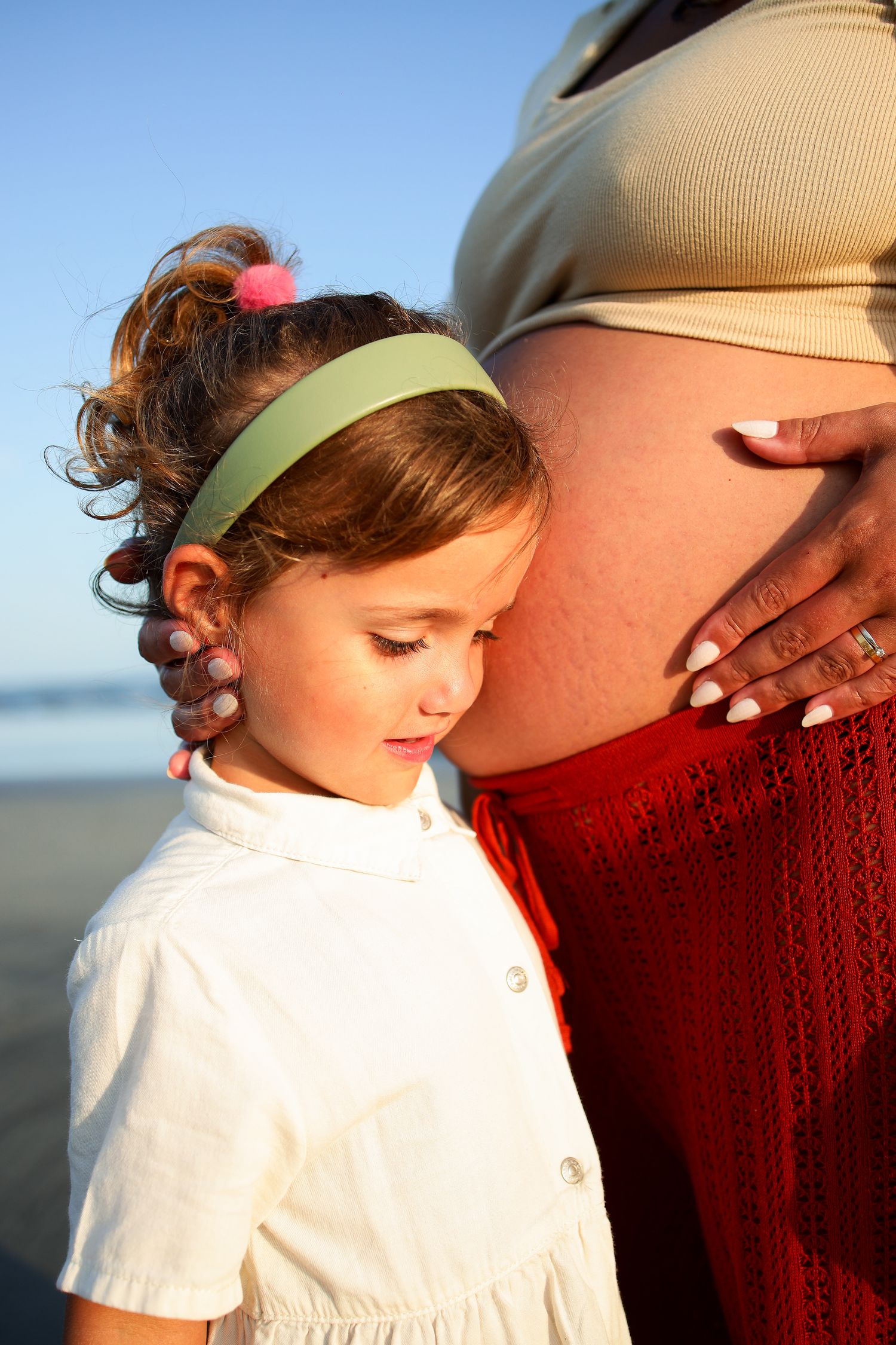 Sweet moment at the beach with a child in white dress and green headband standing next to pregnant belly in warm sunlight.