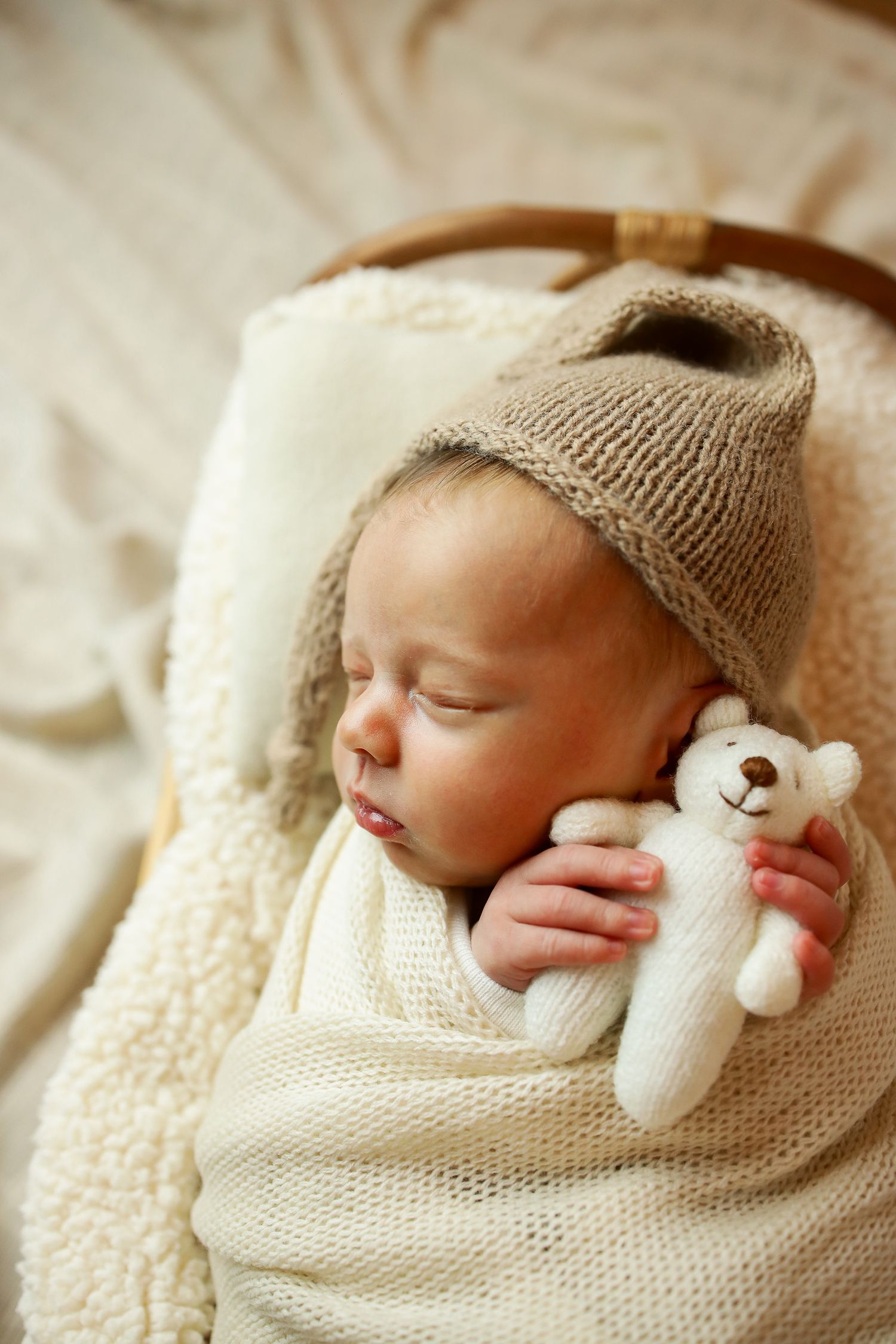 A sleeping baby wrapped in a cream knit blanket holds a small white teddy bear while wearing a beige knitted hat.