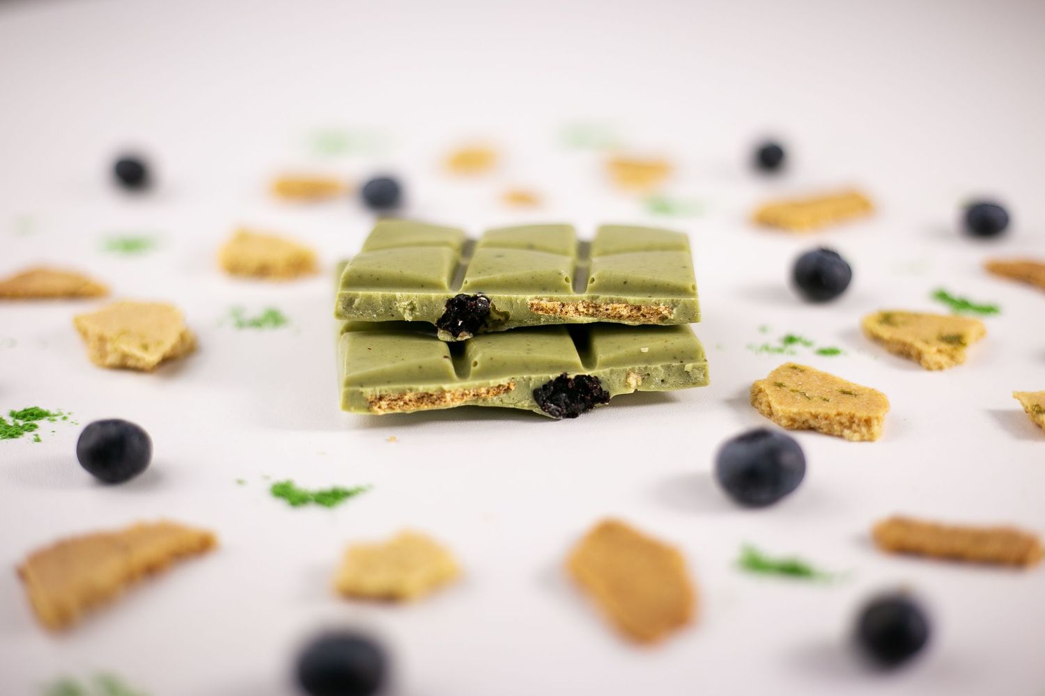 Green matcha chocolate bars stacked with blueberries and graham cracker pieces scattered on white surface.