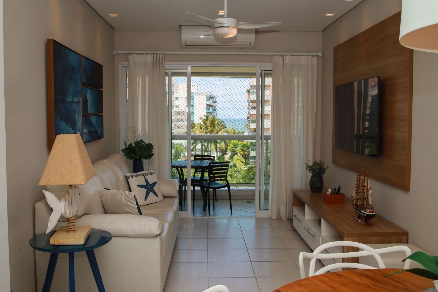Modern coastal apartment living room with ocean view balcony, beige sofa, and mounted TV on wood accent wall.