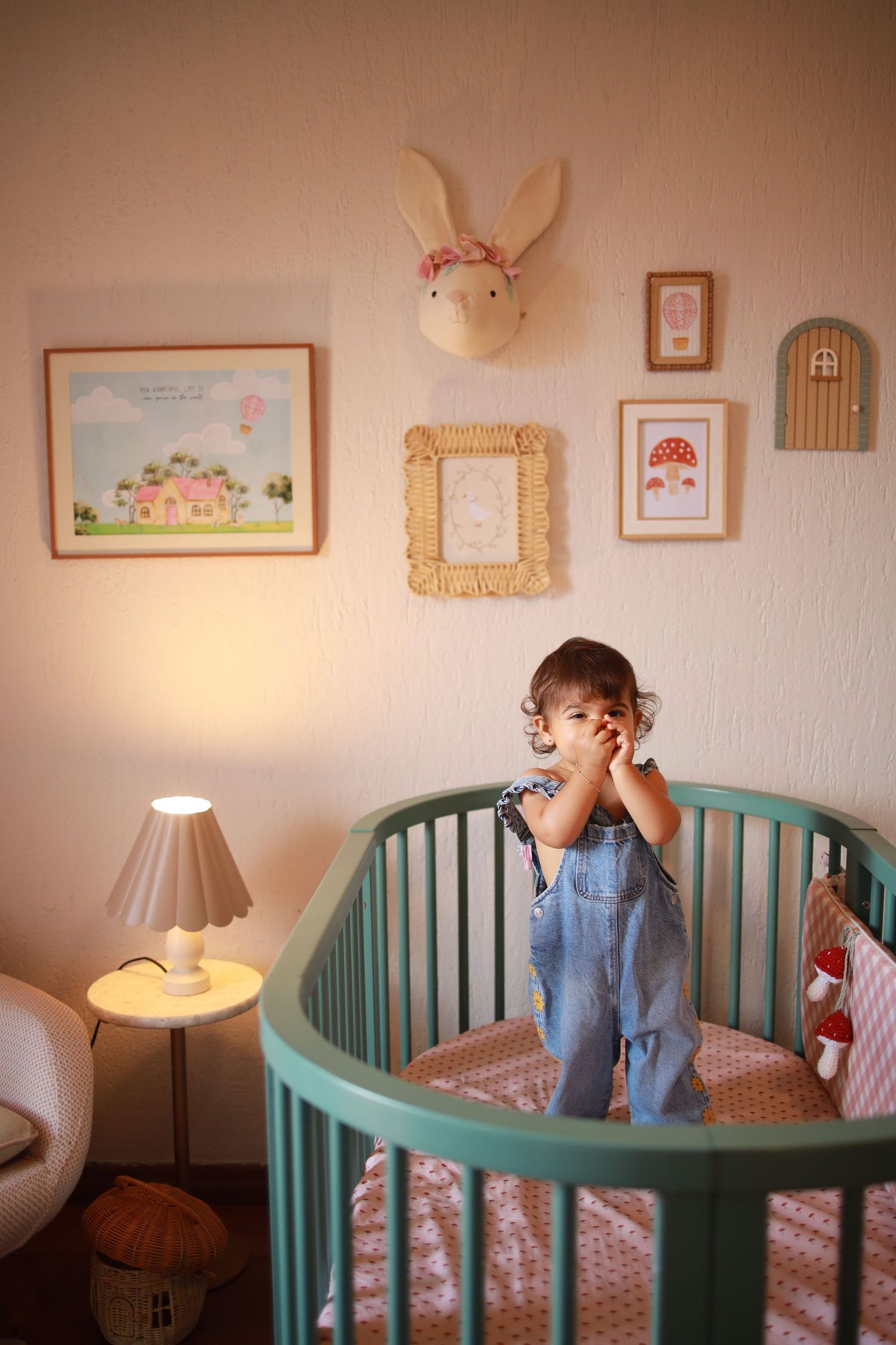 A nursery room with teal crib, bunny wall decor, framed artwork, and soft lamp lighting creating a cozy atmosphere.