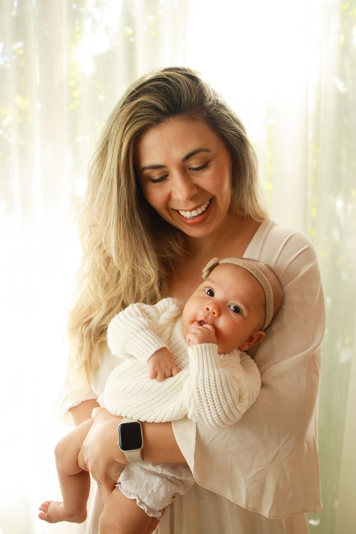 A joyful parent wearing beige holds a baby dressed in a white knit sweater by a sunlit window.