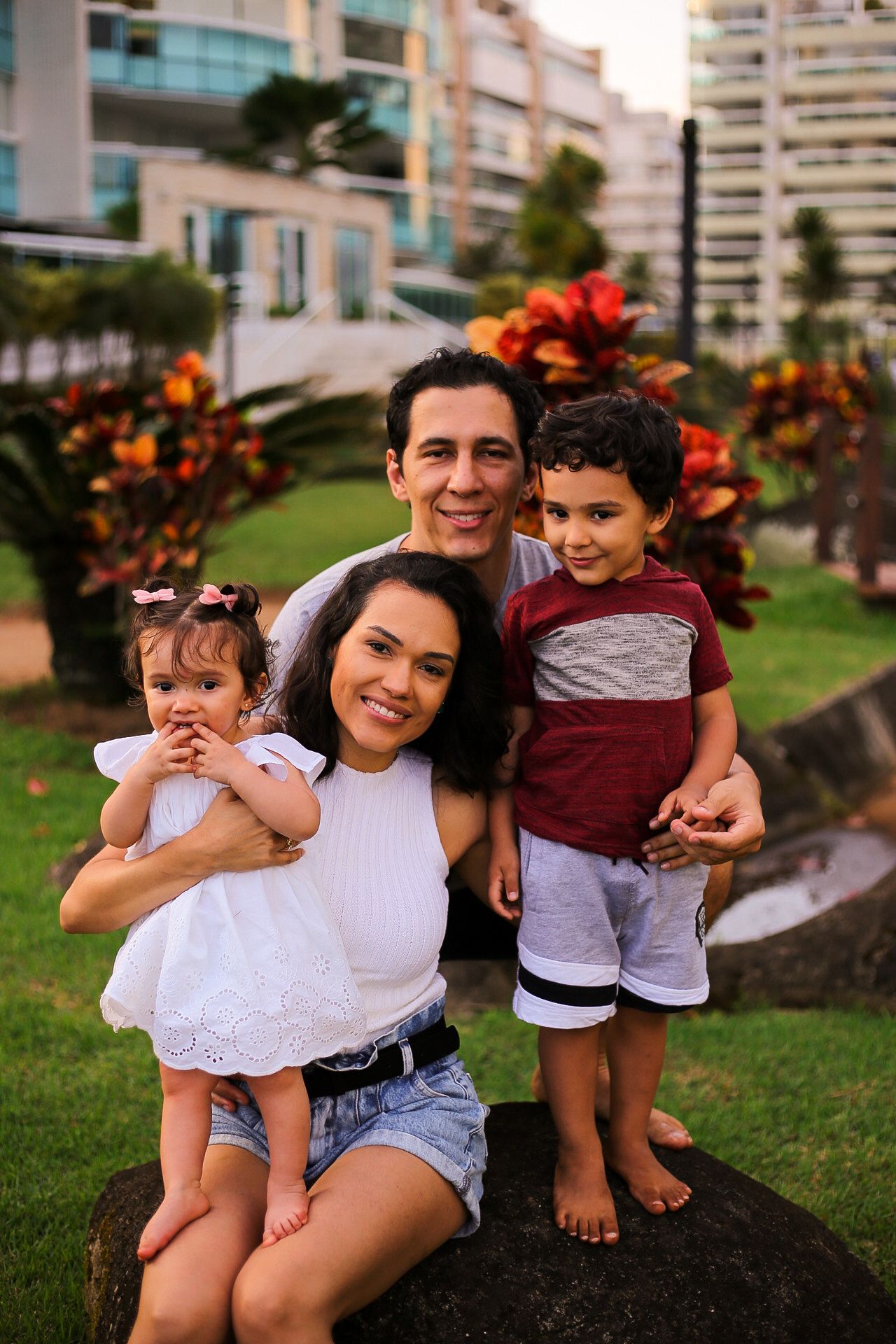 A happy family poses together outdoors on a grassy area with tropical plants and buildings in the background.