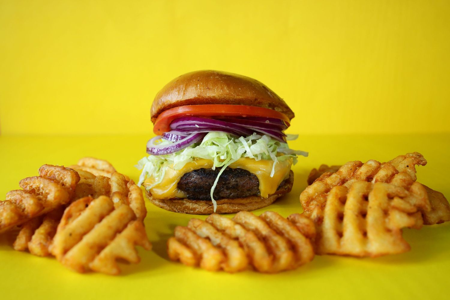 A juicy cheeseburger with fresh toppings surrounded by crispy curly fries on a bright yellow background.
