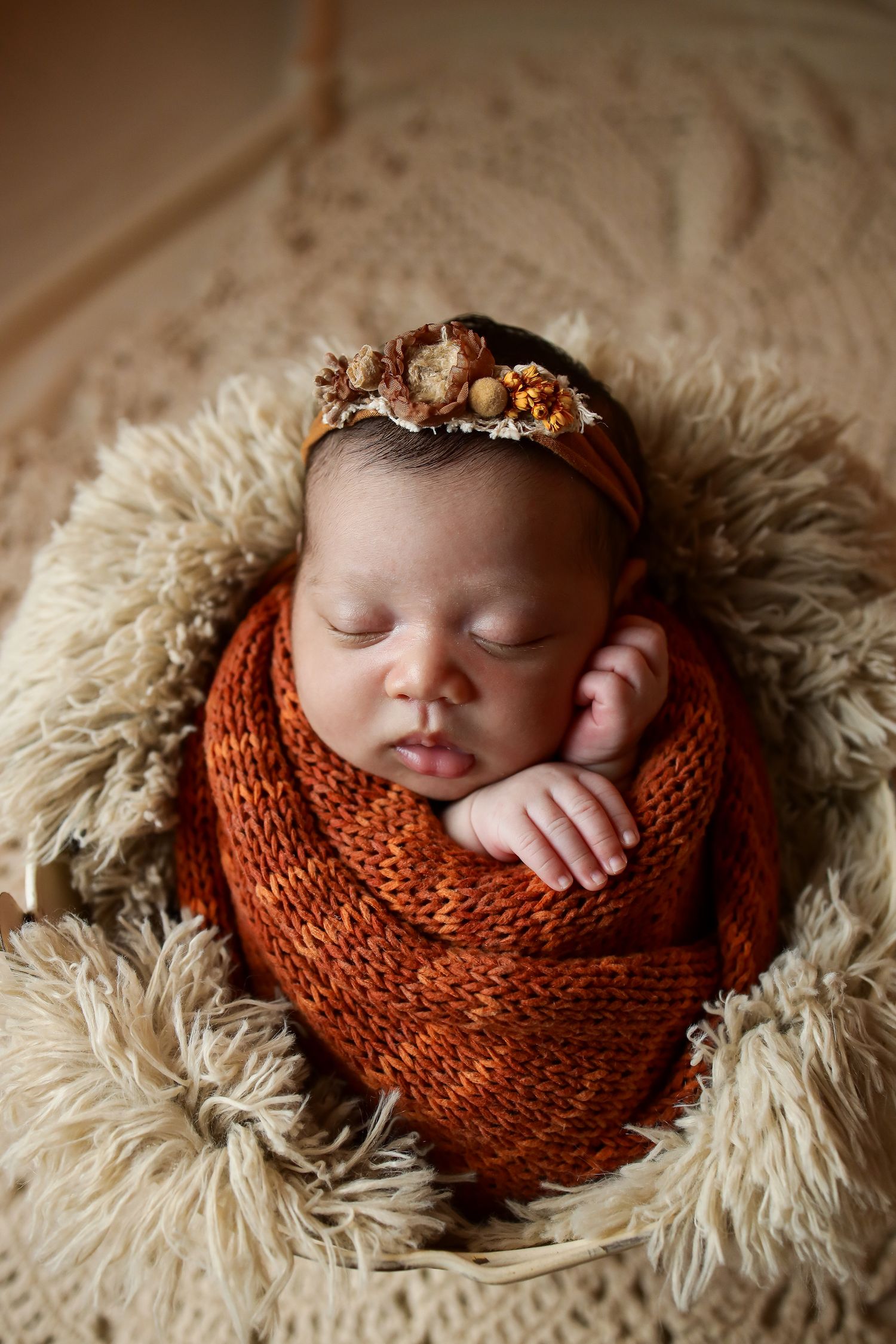 Sleeping newborn wrapped in rustic orange knit swaddle on cream fur blanket with floral headband.