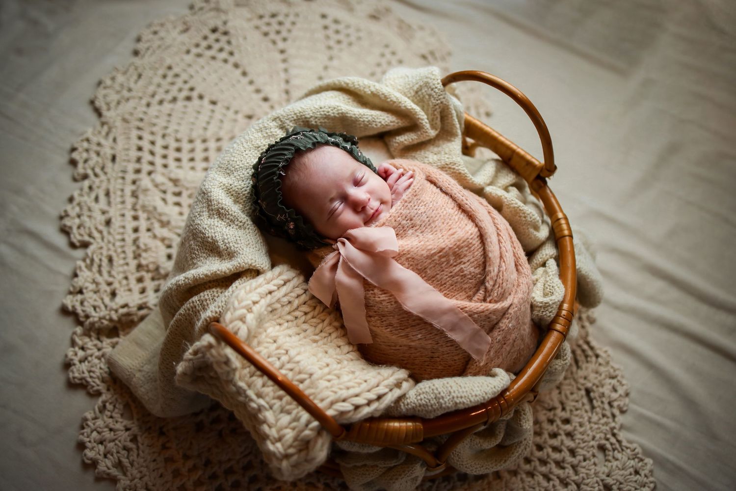Sleepy newborn wrapped in peach swaddle rests peacefully in woven basket on cream lace blanket.