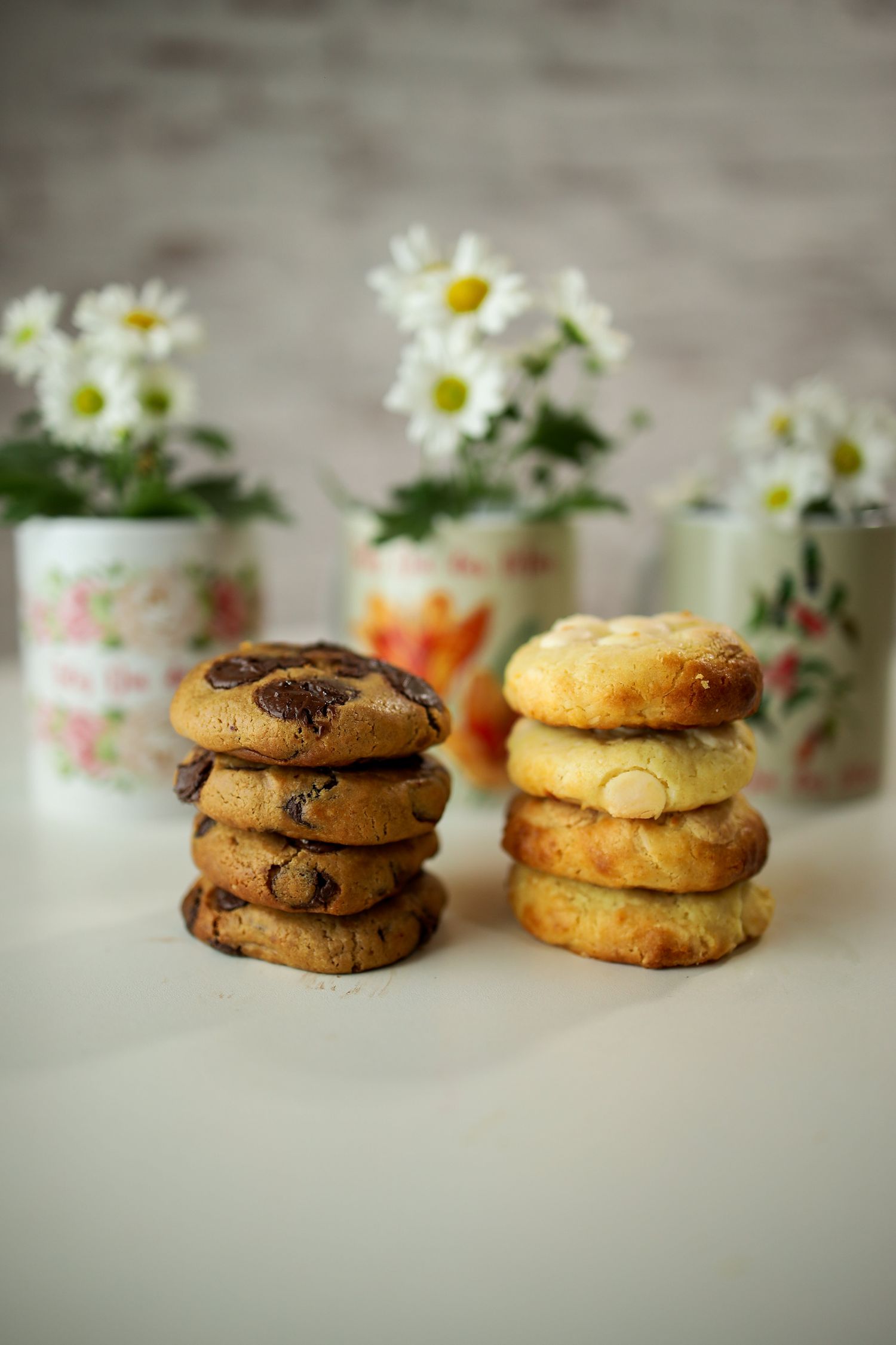 Stacked chocolate chip and sugar cookies with white daisies in vintage floral containers in the background.