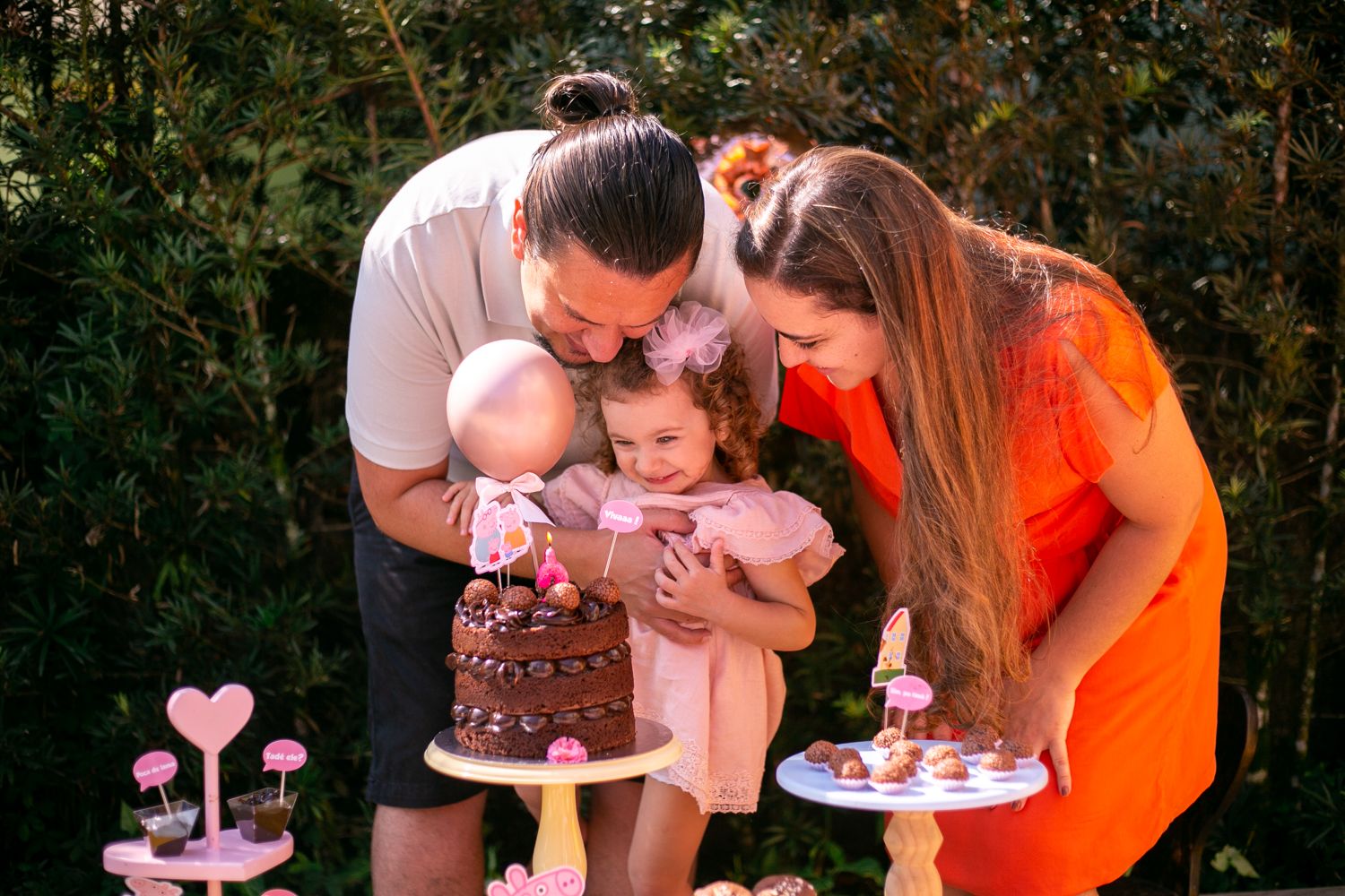 Family celebrates together at outdoor birthday party with chocolate cake and pink decorations on dessert table.