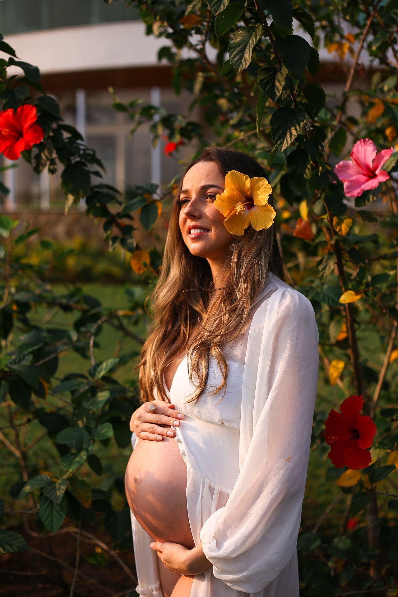 Pregnant figure in a white flowing dress poses in a garden with vibrant hibiscus flowers under golden sunlight.