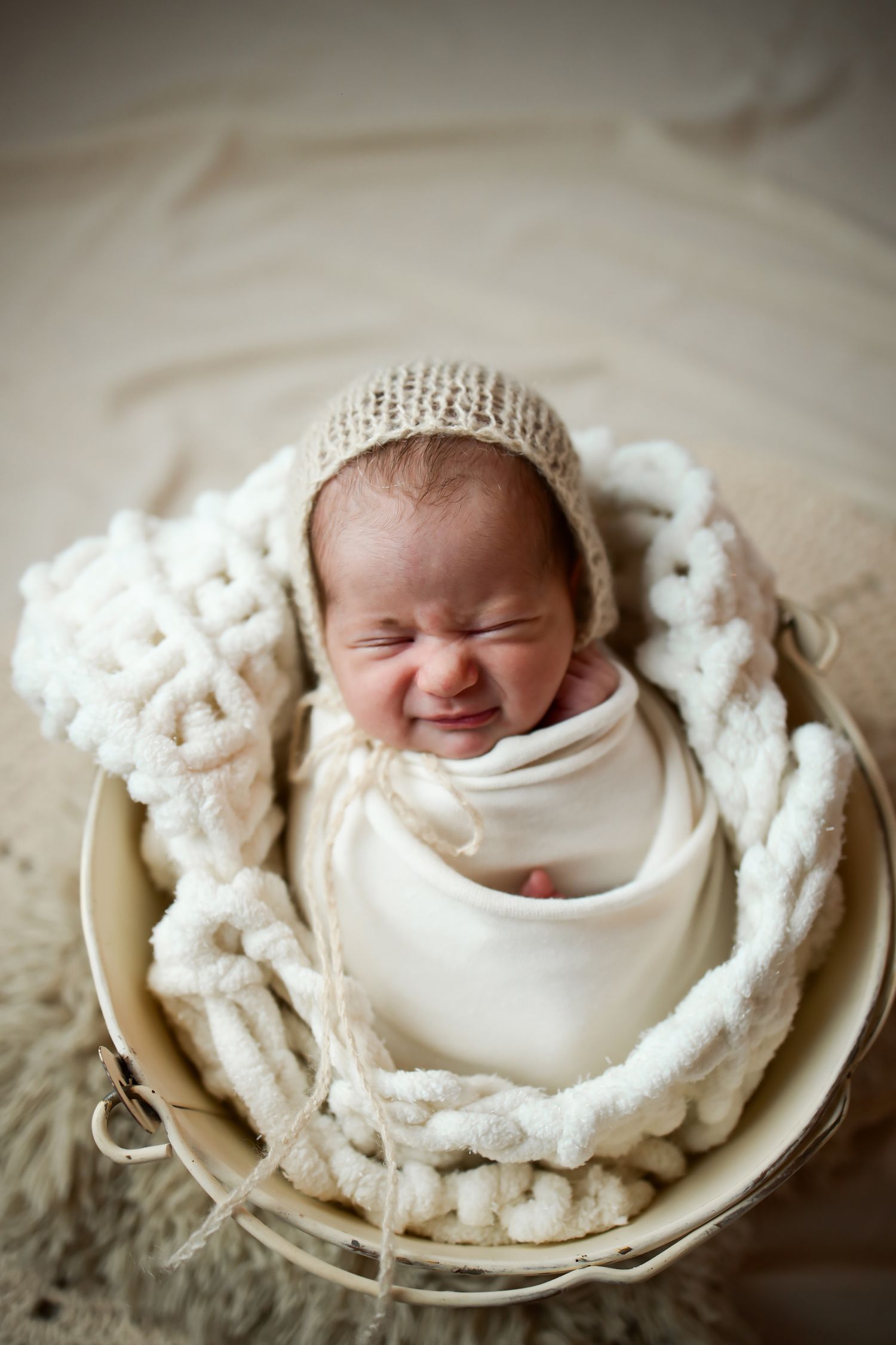 A newborn swaddled in white fabric and wearing a knit bonnet smiles while nestled in a cozy blanket inside a wooden bowl.