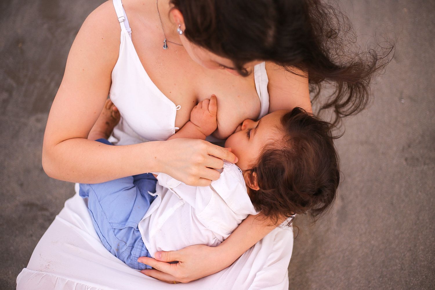 A mother in white top gently breastfeeding her baby while sitting on a gray background.