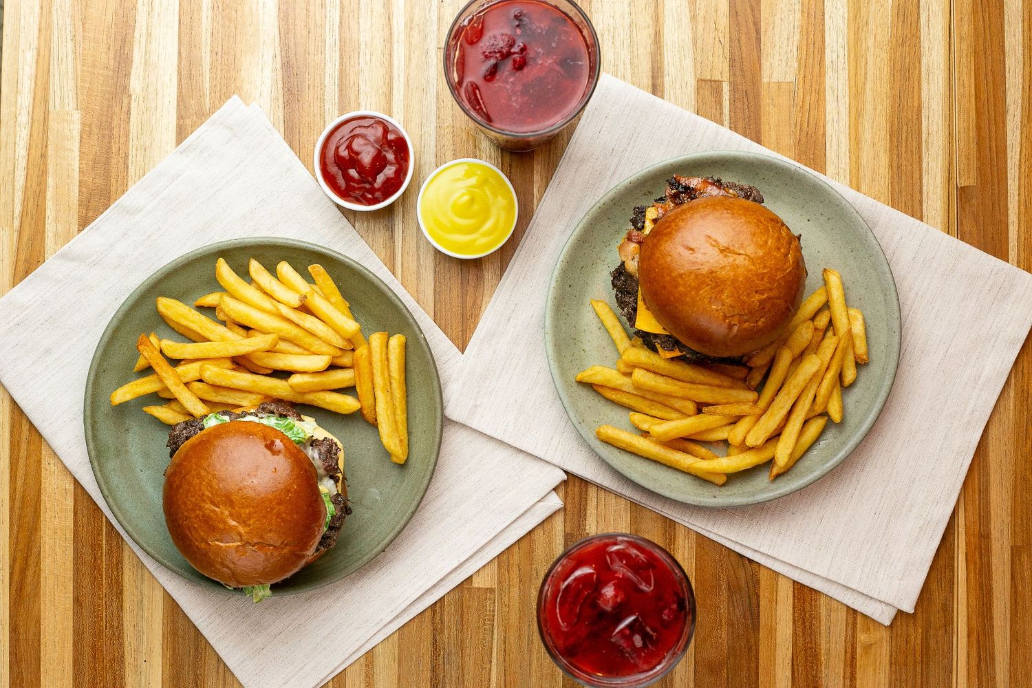 Two classic cheeseburgers served with crispy french fries and assorted condiment sauces on gray plates and butcher paper.