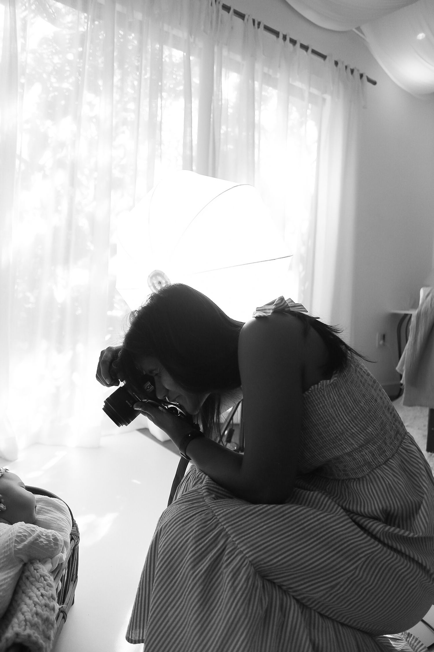A silhouetted figure in a striped dress crouches by a window with a camera, taking photos in black and white.