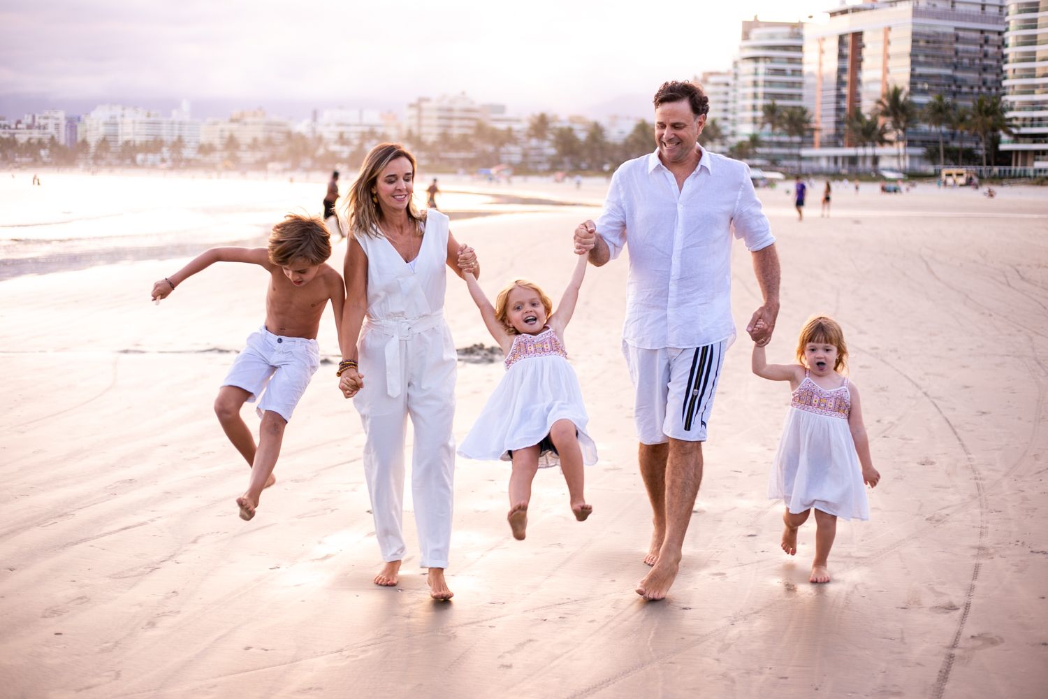A family dressed in white outfits plays together and walks along a sunny beach shoreline.