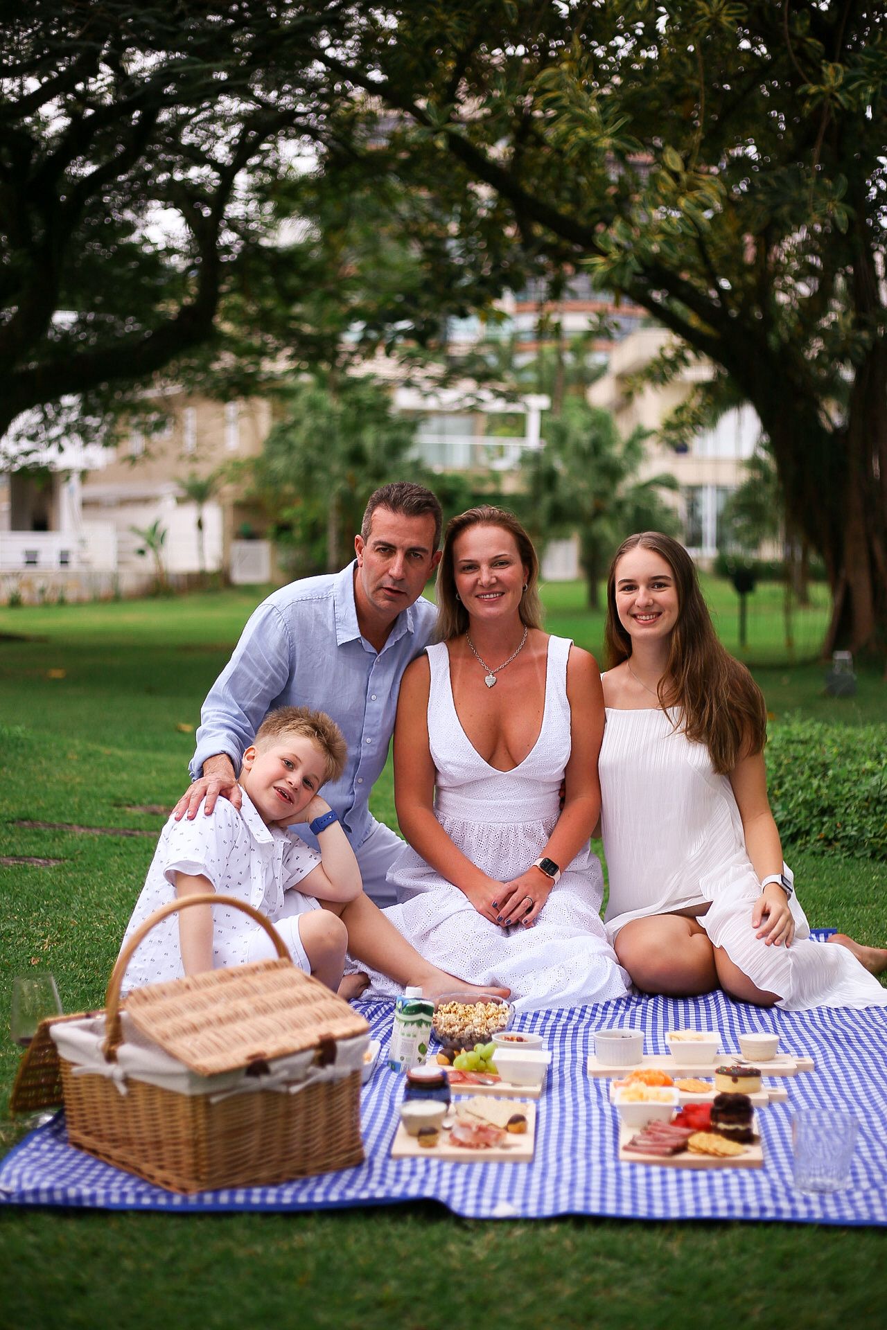 A happy family enjoying a summer picnic on a blue checkered blanket with a wicker basket and food spread outdoors.