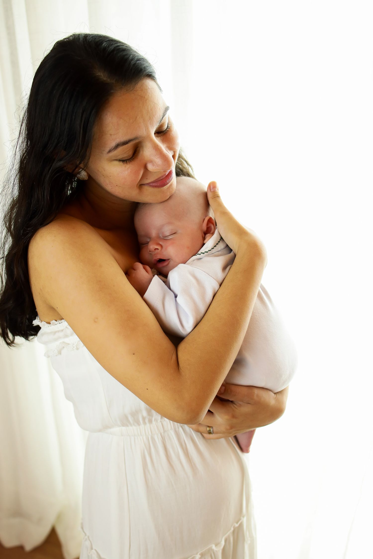 A loving mother in a white dress holds and snuggles with her sleeping newborn baby in a tender moment against bright light.