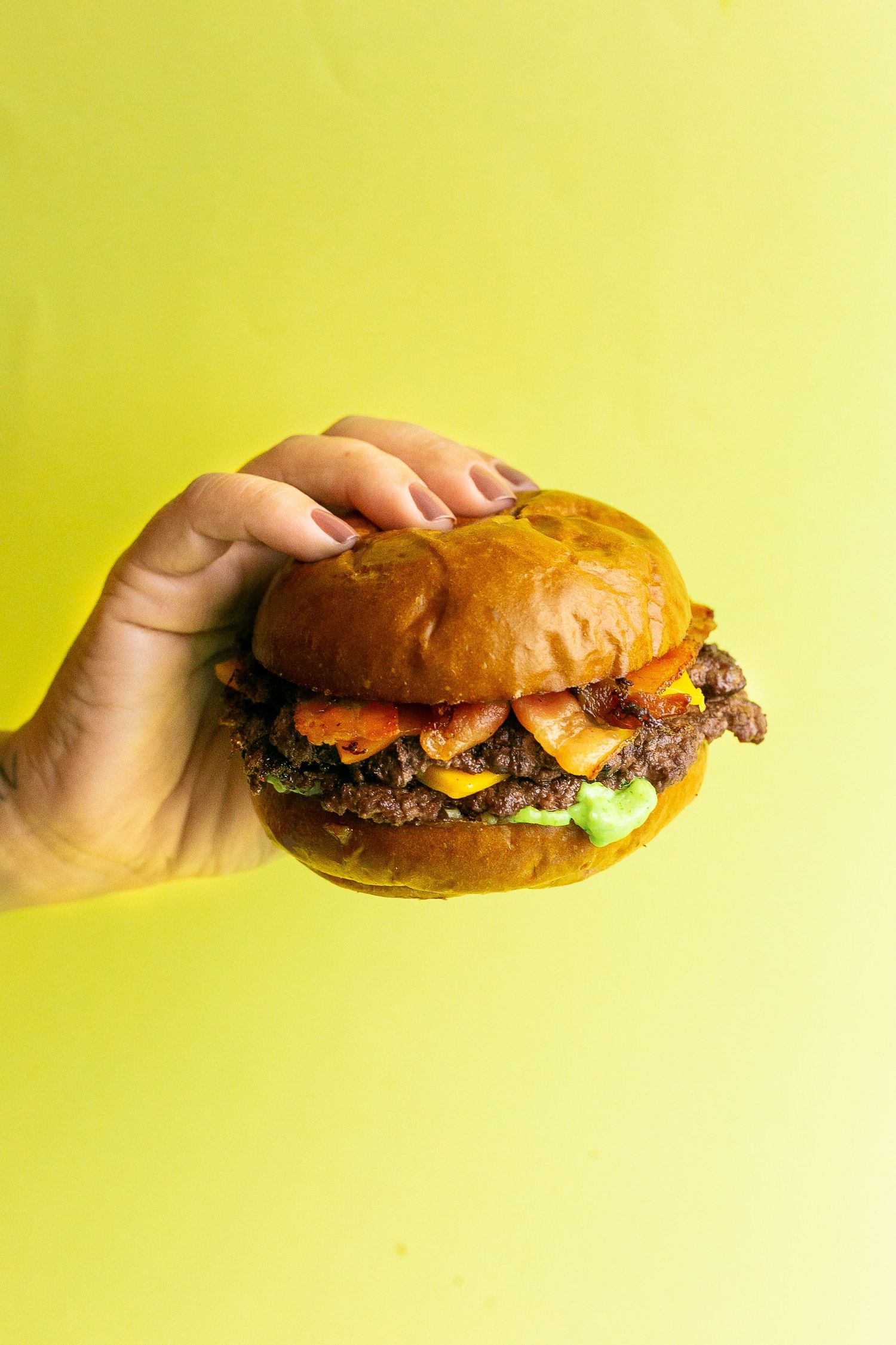Hand holding a juicy cheeseburger with lettuce and bacon on a brioche bun against a yellow background.