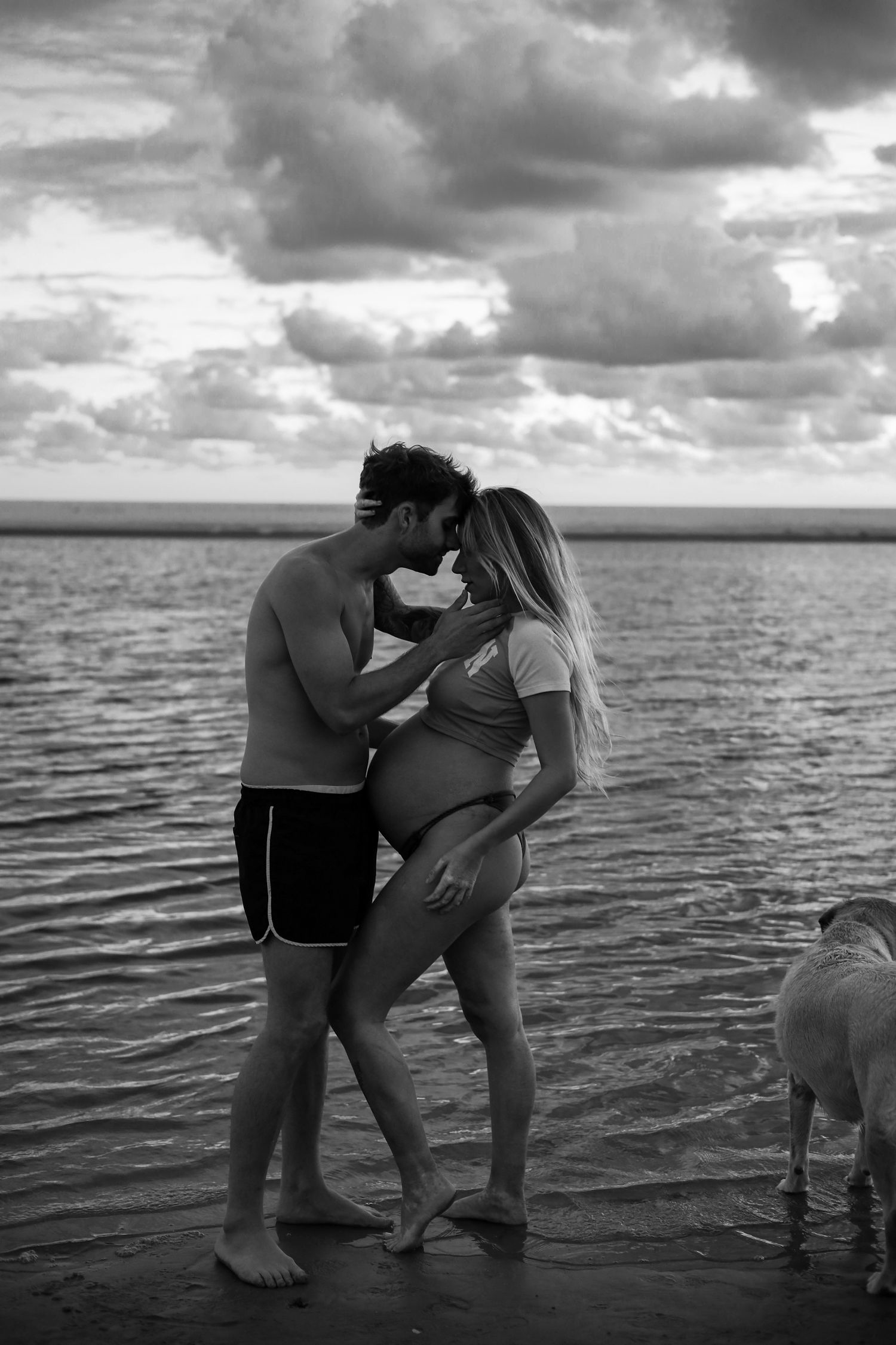 Silhouetted couple embraces on beach at sunset with dramatic cloudy sky.