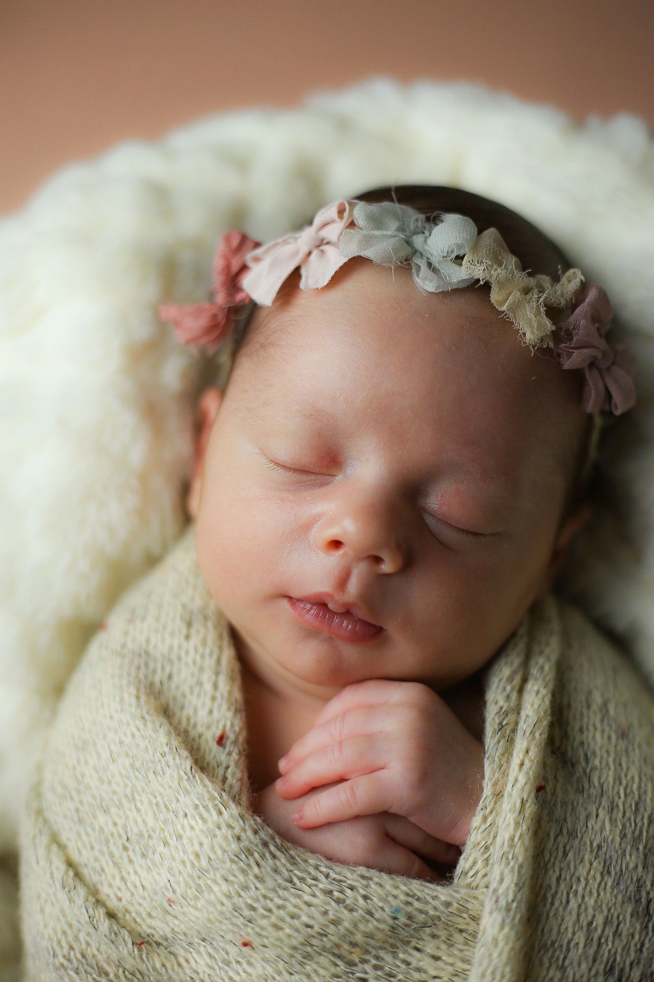 Sleeping newborn peacefully swaddled in cream knit wrap wearing delicate floral headband on soft white blanket.