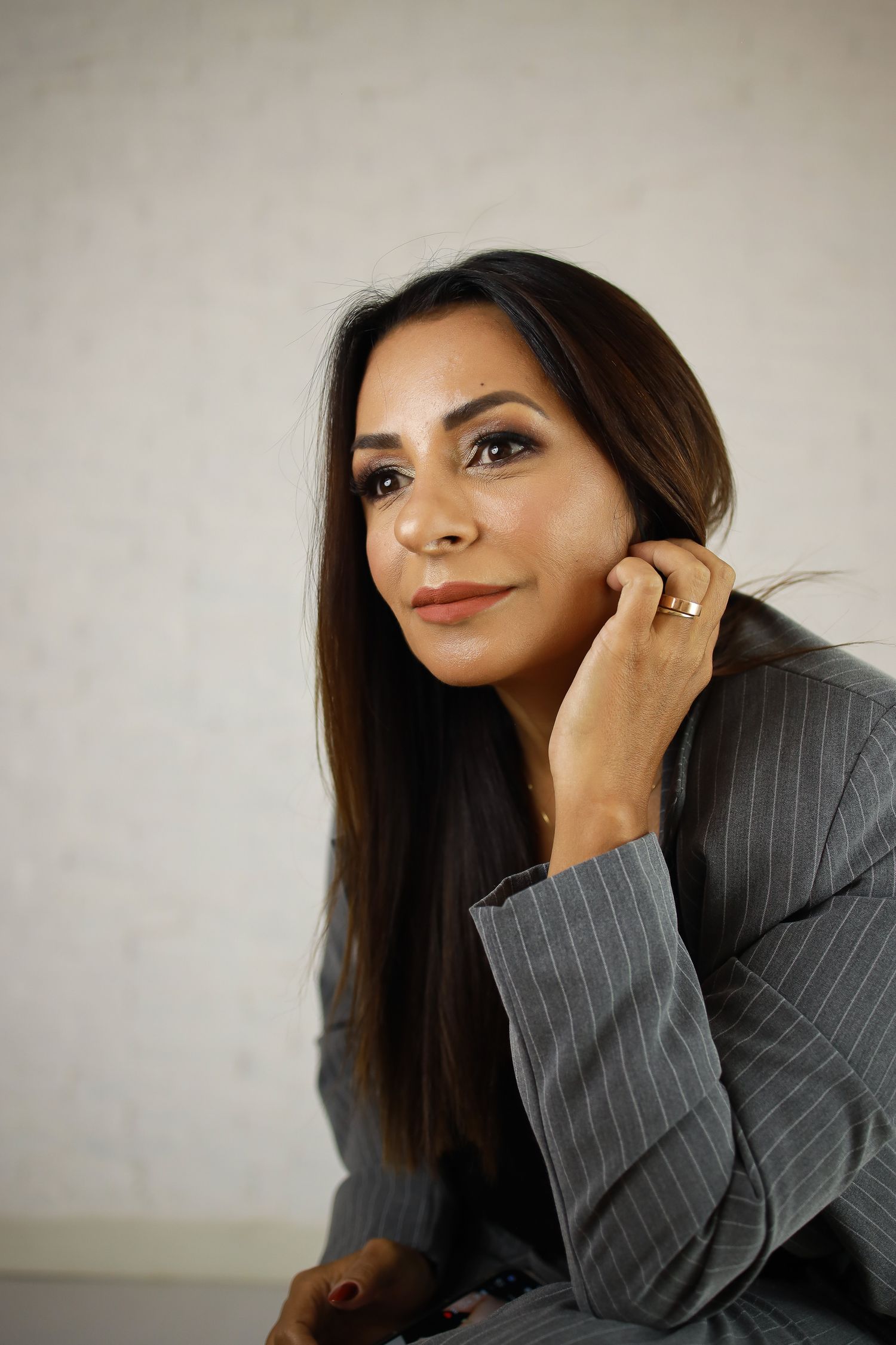 Professional portrait of businesswoman in pinstripe suit jacket posing with hand near face against neutral background.