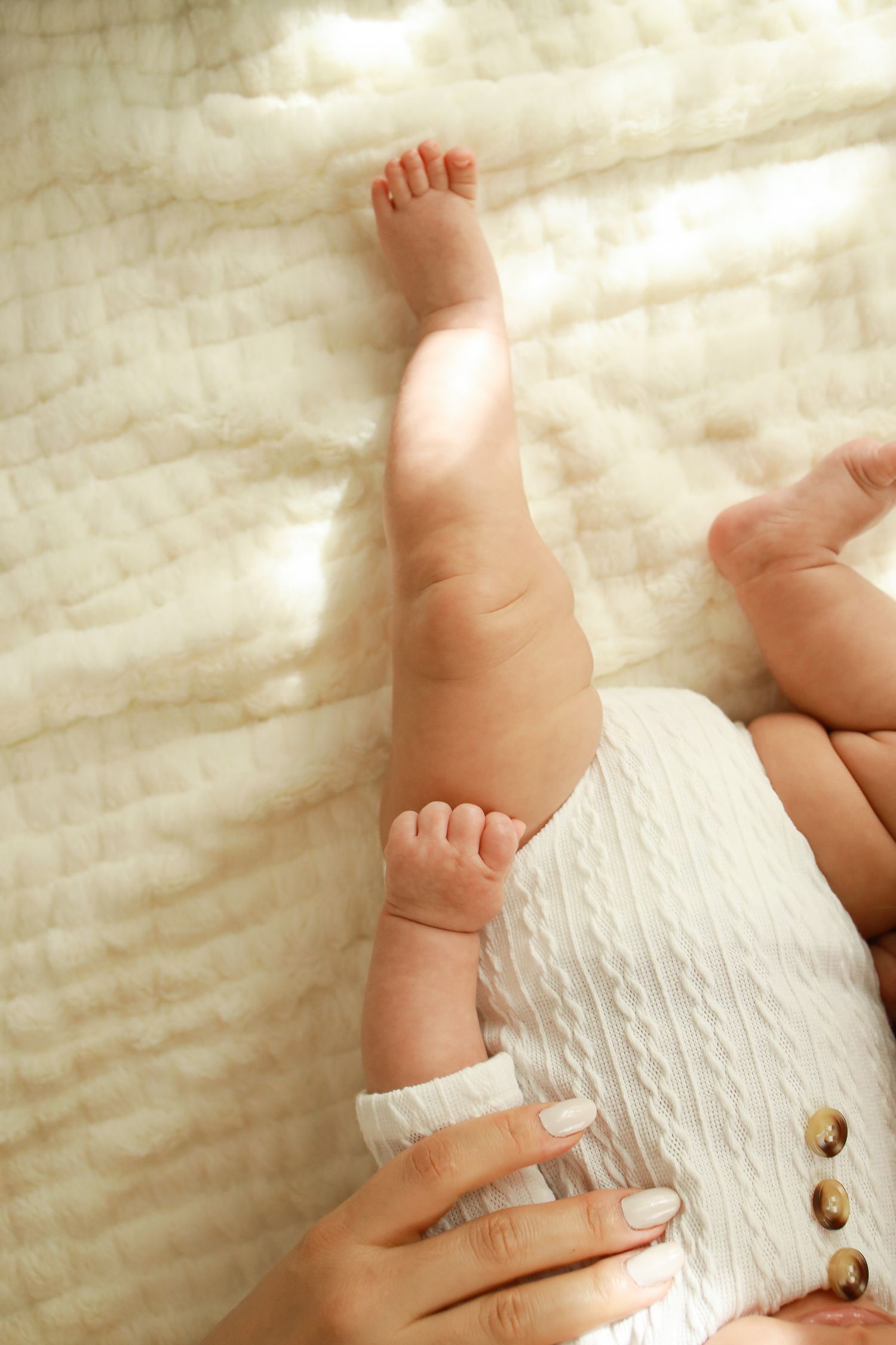 A baby wearing a white textured romper with gold buttons lies on cream-colored bedding as adult hands adjust the clothing.