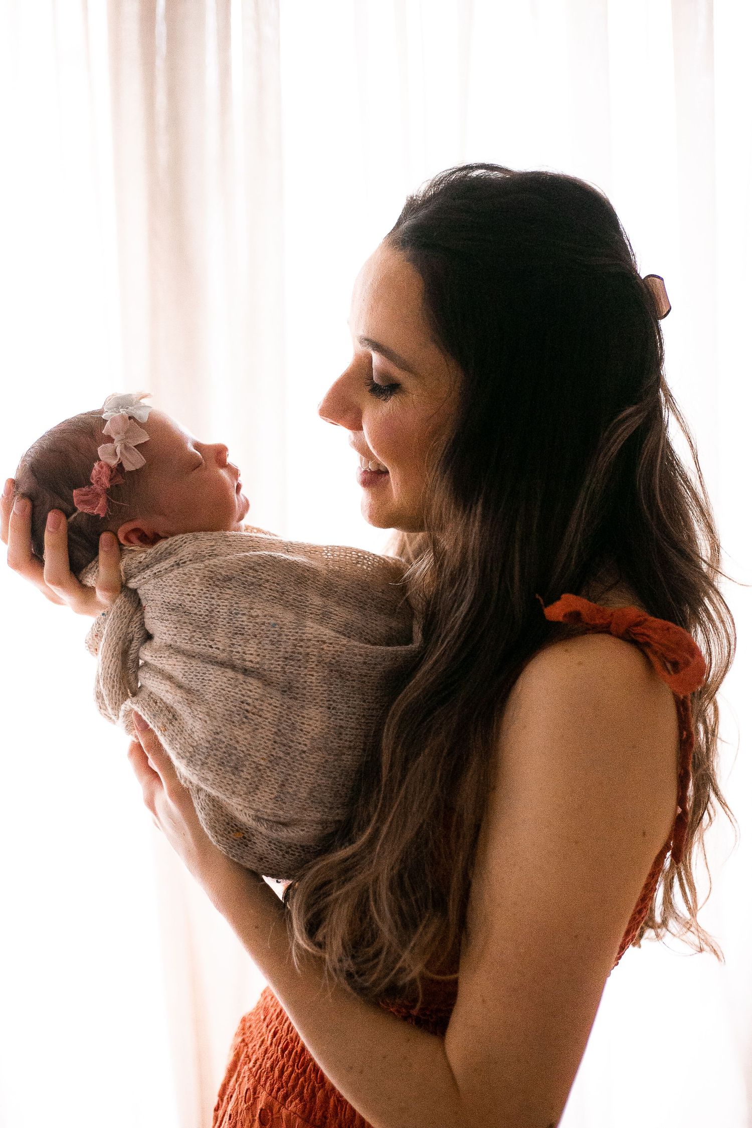 Mother in orange dress lovingly holds newborn baby swaddled in beige wrap against soft window light in tender moment.