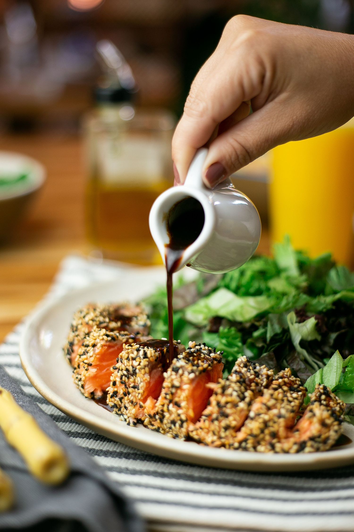 Sesame crusted salmon fillets with fresh greens being drizzled with dark sauce on a white plate.