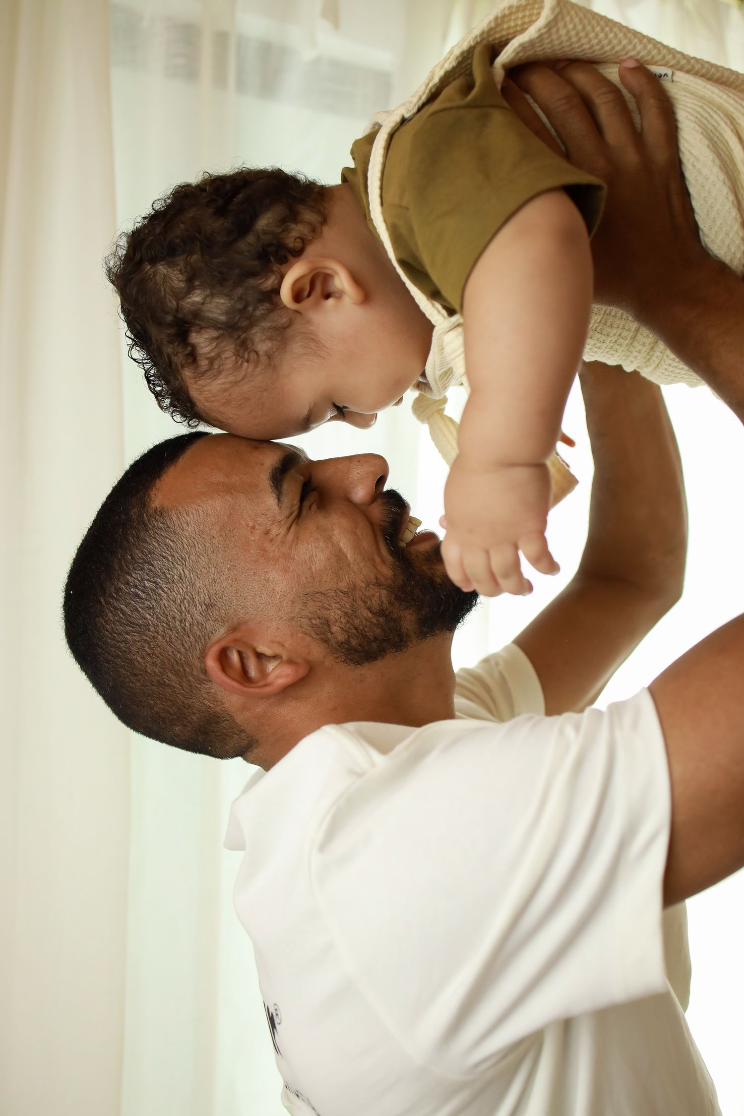 A loving parent shares a tender moment holding up their baby against a soft curtained background.