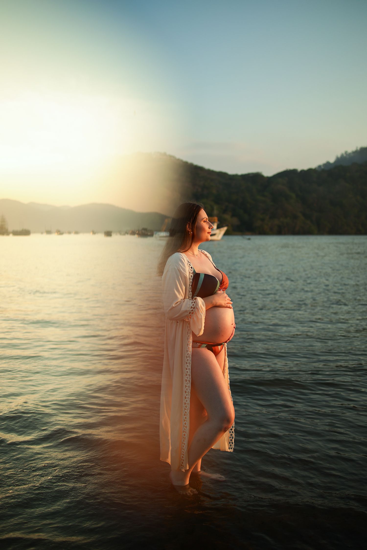 A pregnant woman stands in water at sunset wearing a white robe against a mountain lake backdrop.