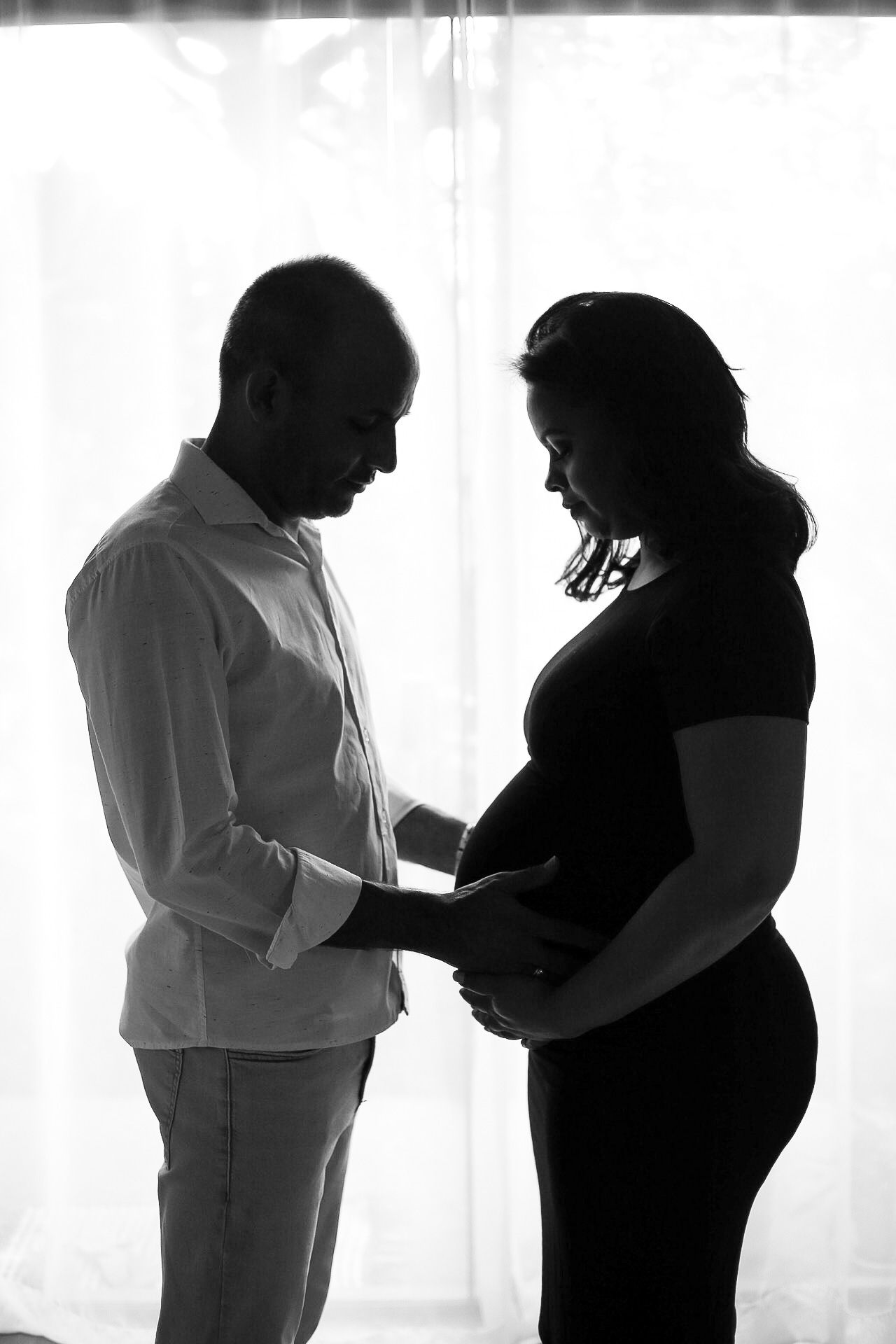 A silhouetted couple shares an intimate moment as they touch a pregnant belly in front of a backlit window.
