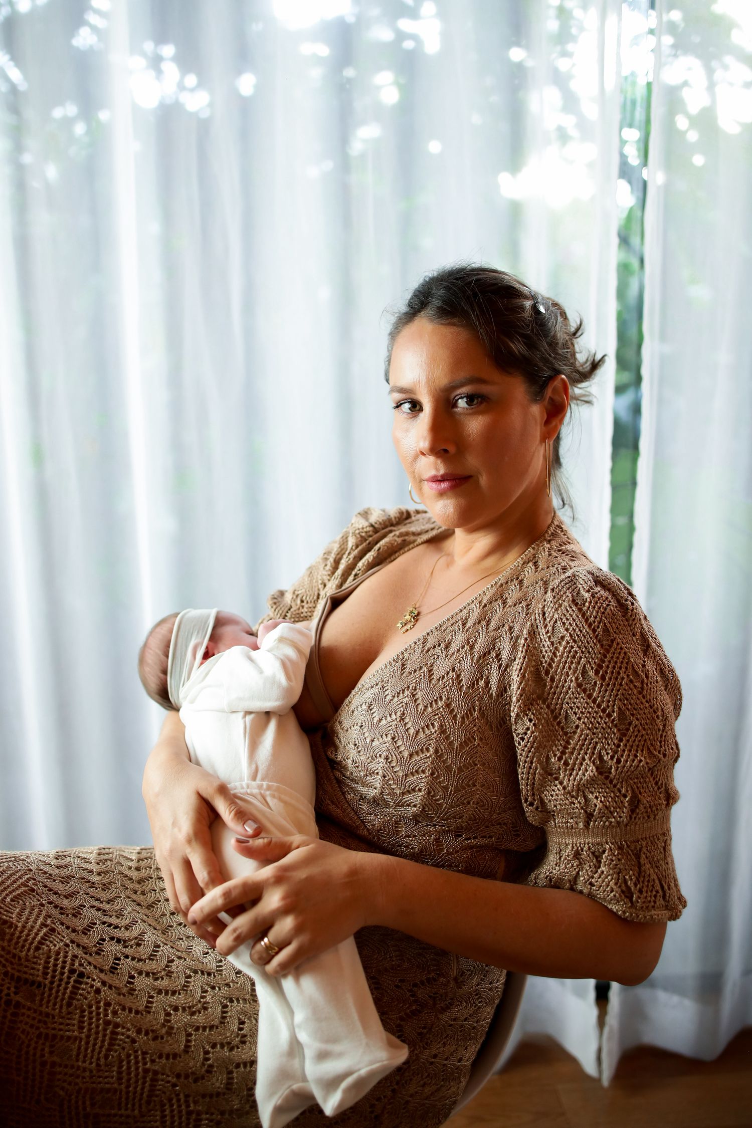 A person wearing a beige cable knit sweater holds a newborn baby wrapped in white fabric near a window with sheer curtains.