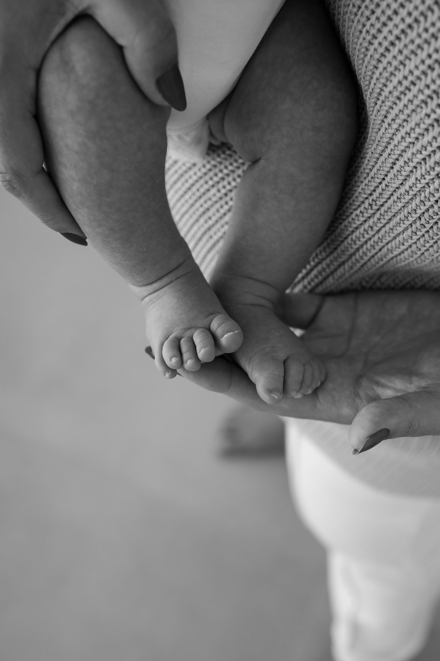 Close-up black and white photo of tiny baby feet being gently held in adult hands with a white knit blanket.