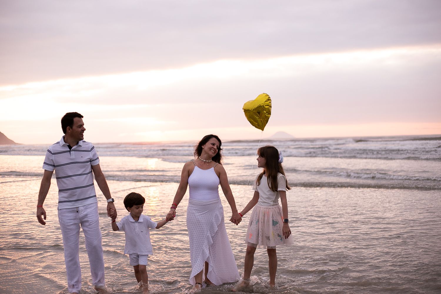 Family walking along beach at sunset holding yellow heart balloon with ocean waves in background.