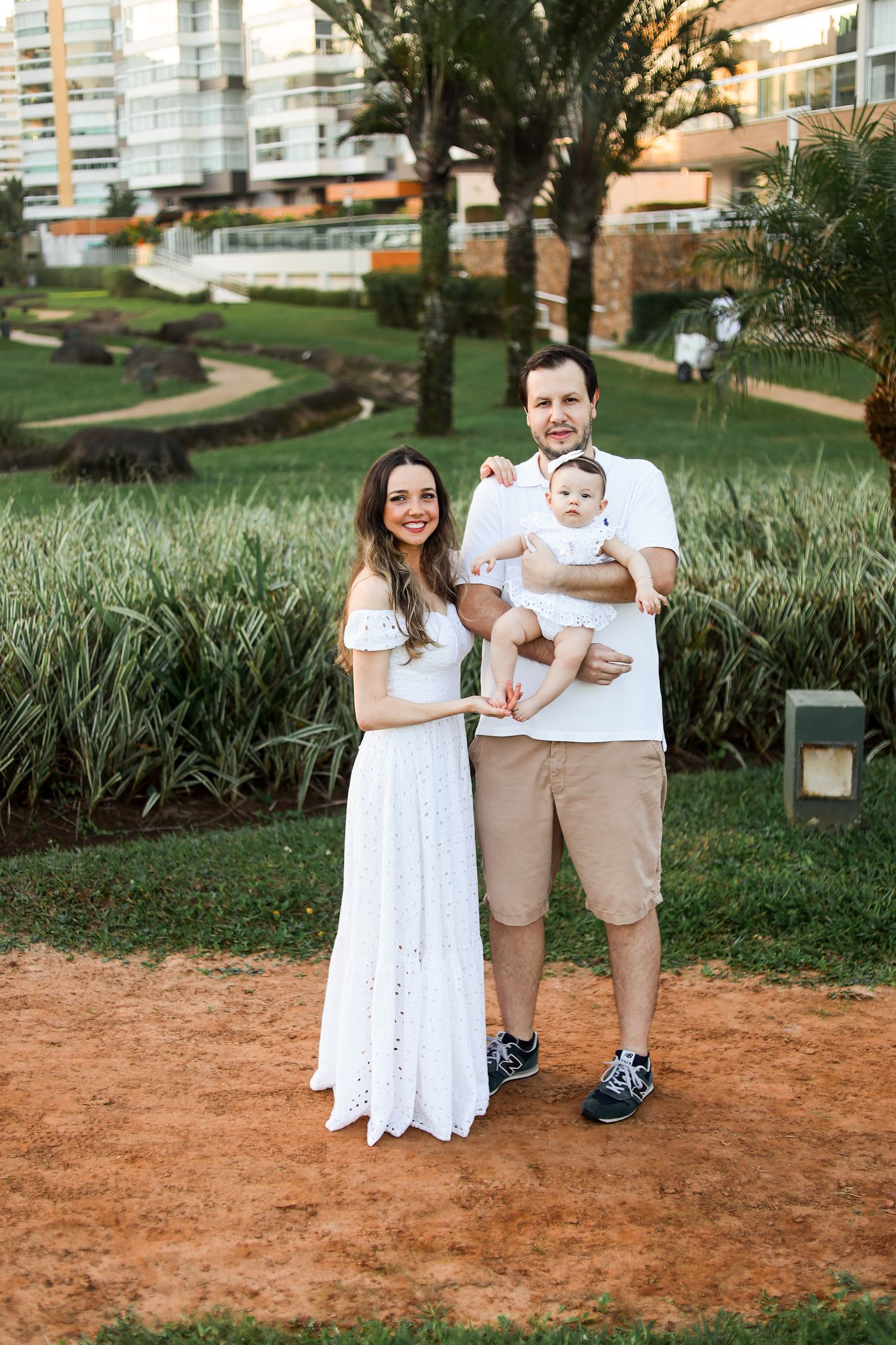 Family wearing coordinated white and beige outfits poses playfully together on a dirt path near landscaped gardens.