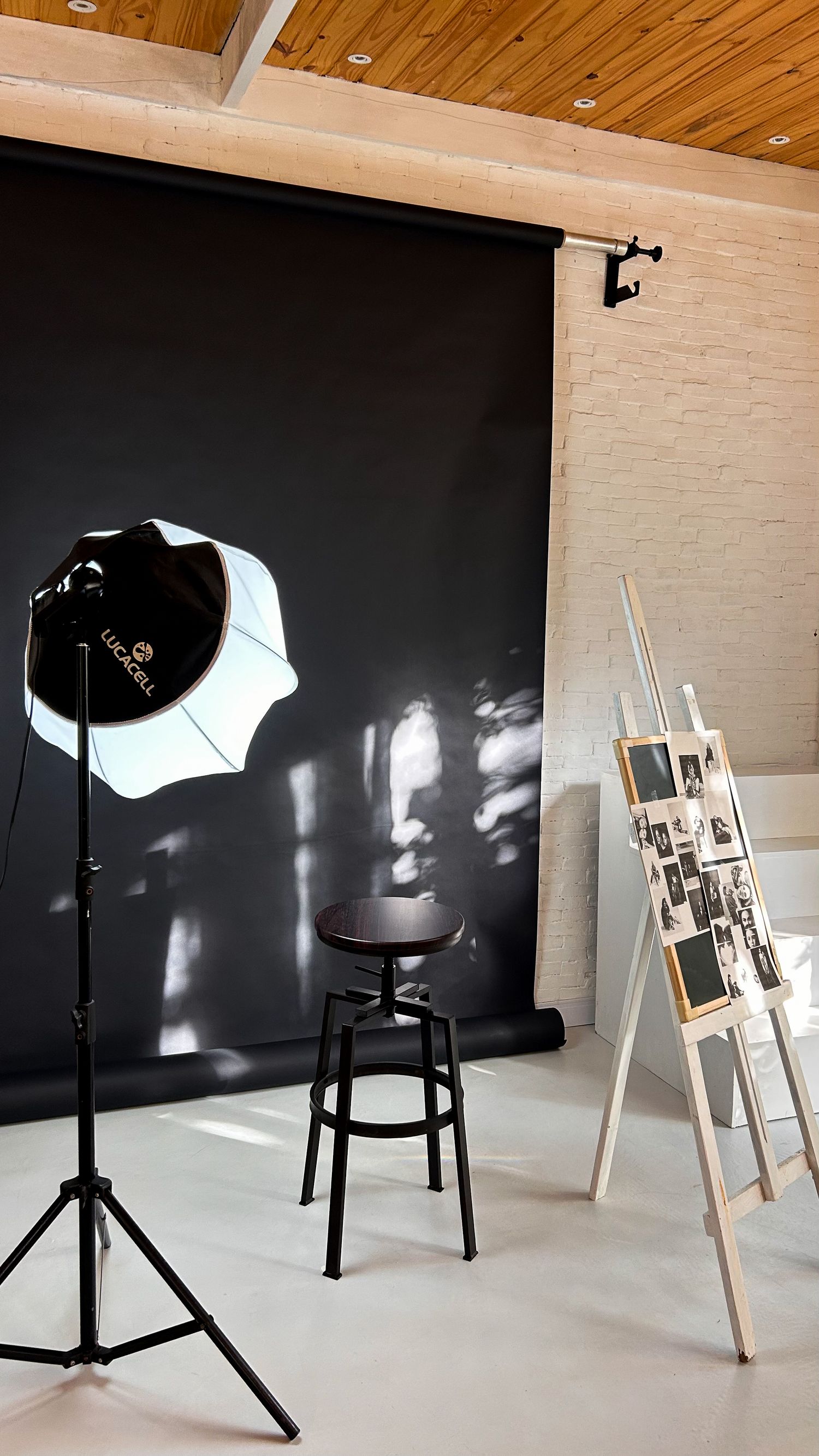 Professional photography studio with black backdrop, umbrella light on tripod, stool, and white easel displaying framed photos.