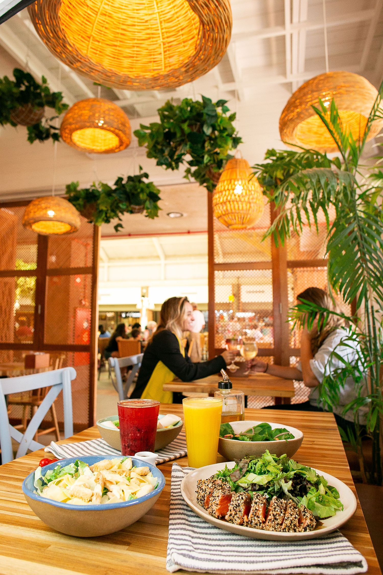 Trendy restaurant interior with wicker pendant lights, tropical plants and colorful healthy food bowls on wooden table.