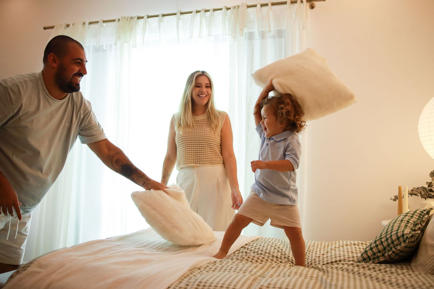 A family shares a playful pillow fight moment together on a bed in a sunlit bedroom.
