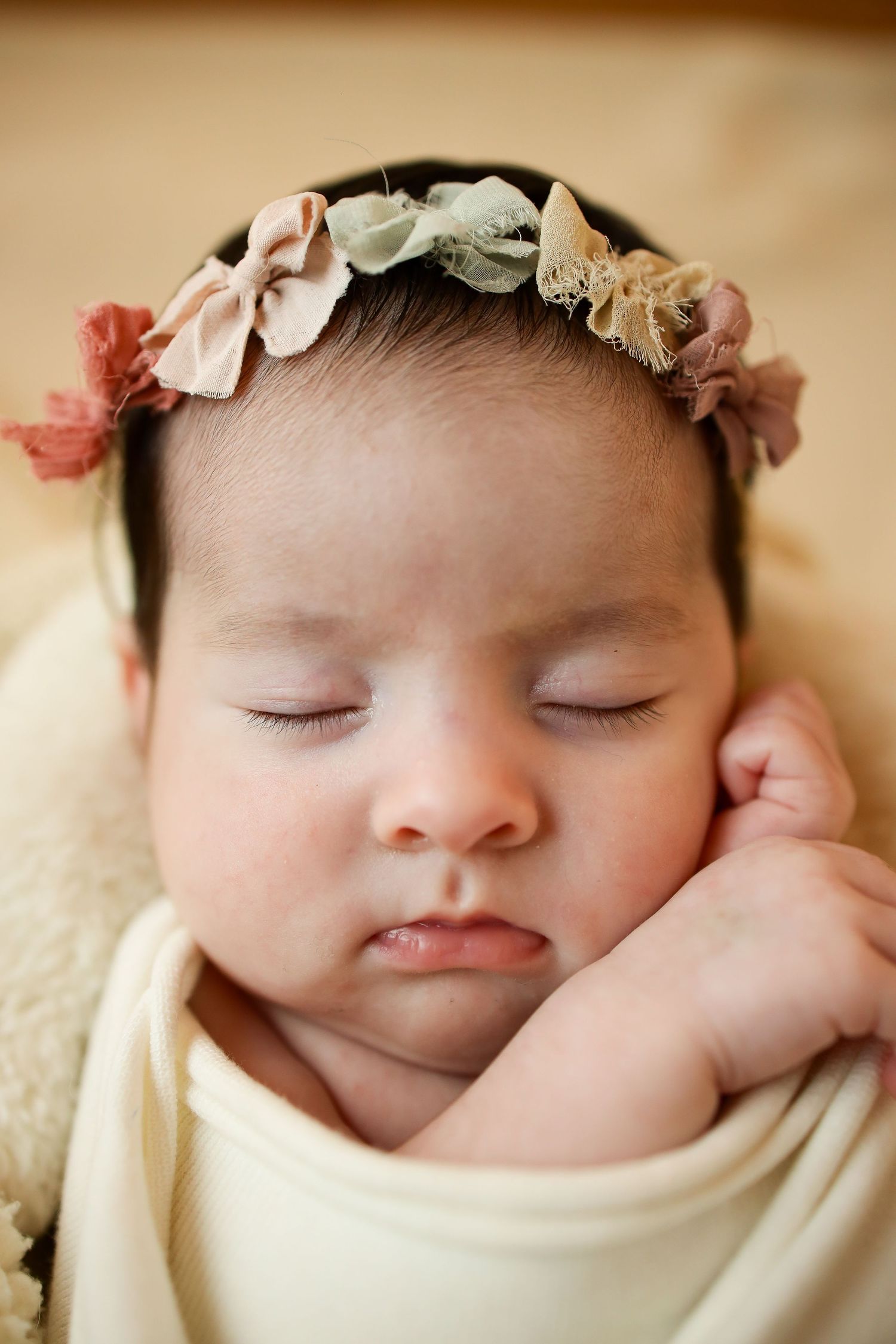 A sleeping newborn wearing a delicate floral headband is wrapped in a soft cream-colored blanket.